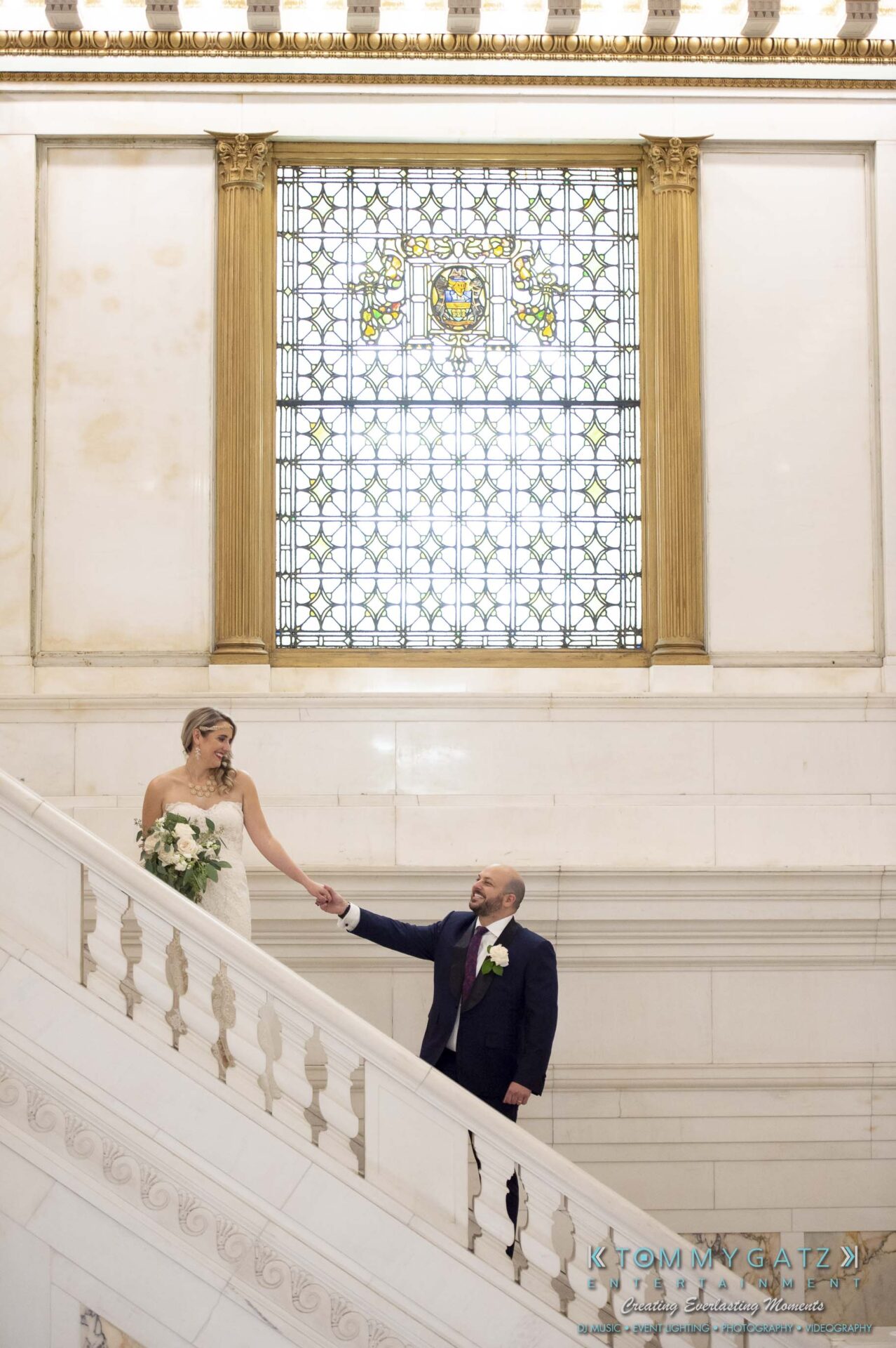 A bride and groom hold hands while ascending a grand marble staircase, with a large stained glass window in the background.