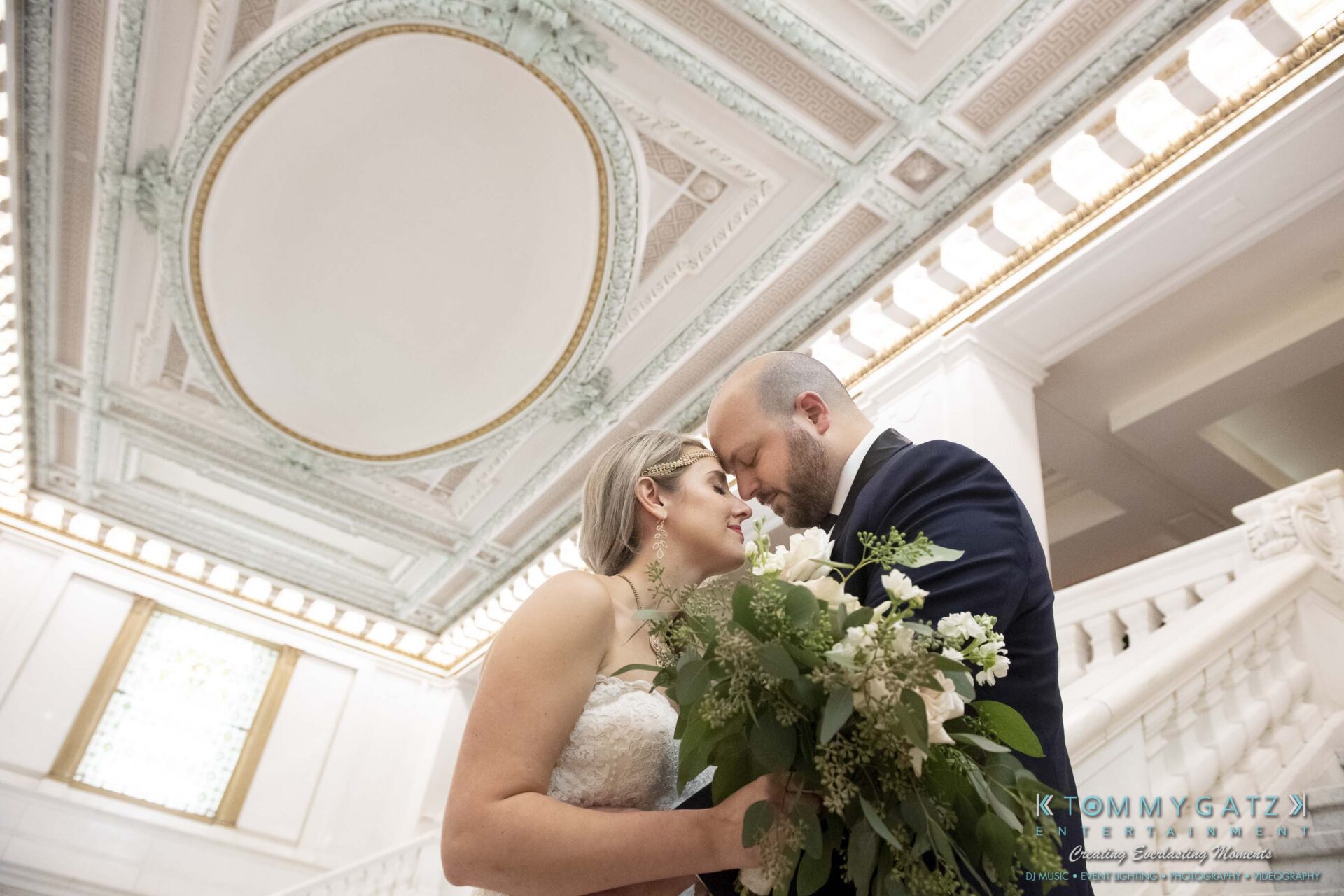 A couple in formal attire embraces holding a bouquet, standing on a staircase in an elegant building with ornate ceilings.