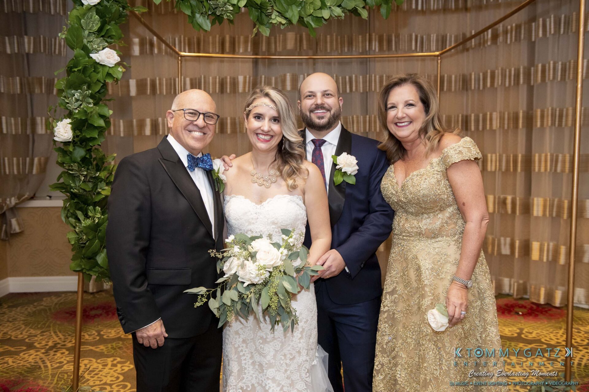 A bride and groom stand with an older man and woman in formal attire, posing in front of a floral arch at an indoor event.