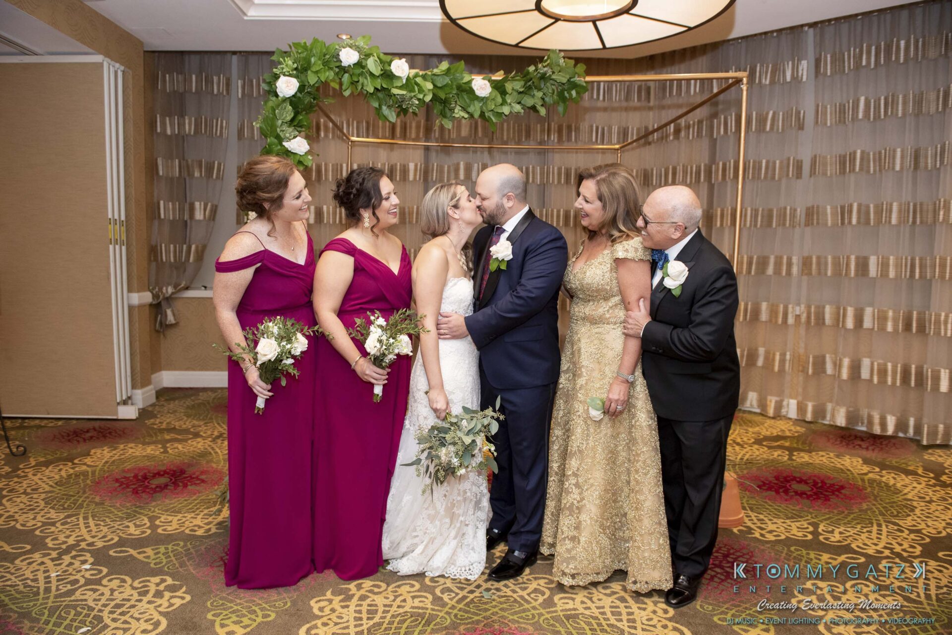 A bride and groom kiss, surrounded by four people in formal attire, standing in front of a floral arch in a decorated room.