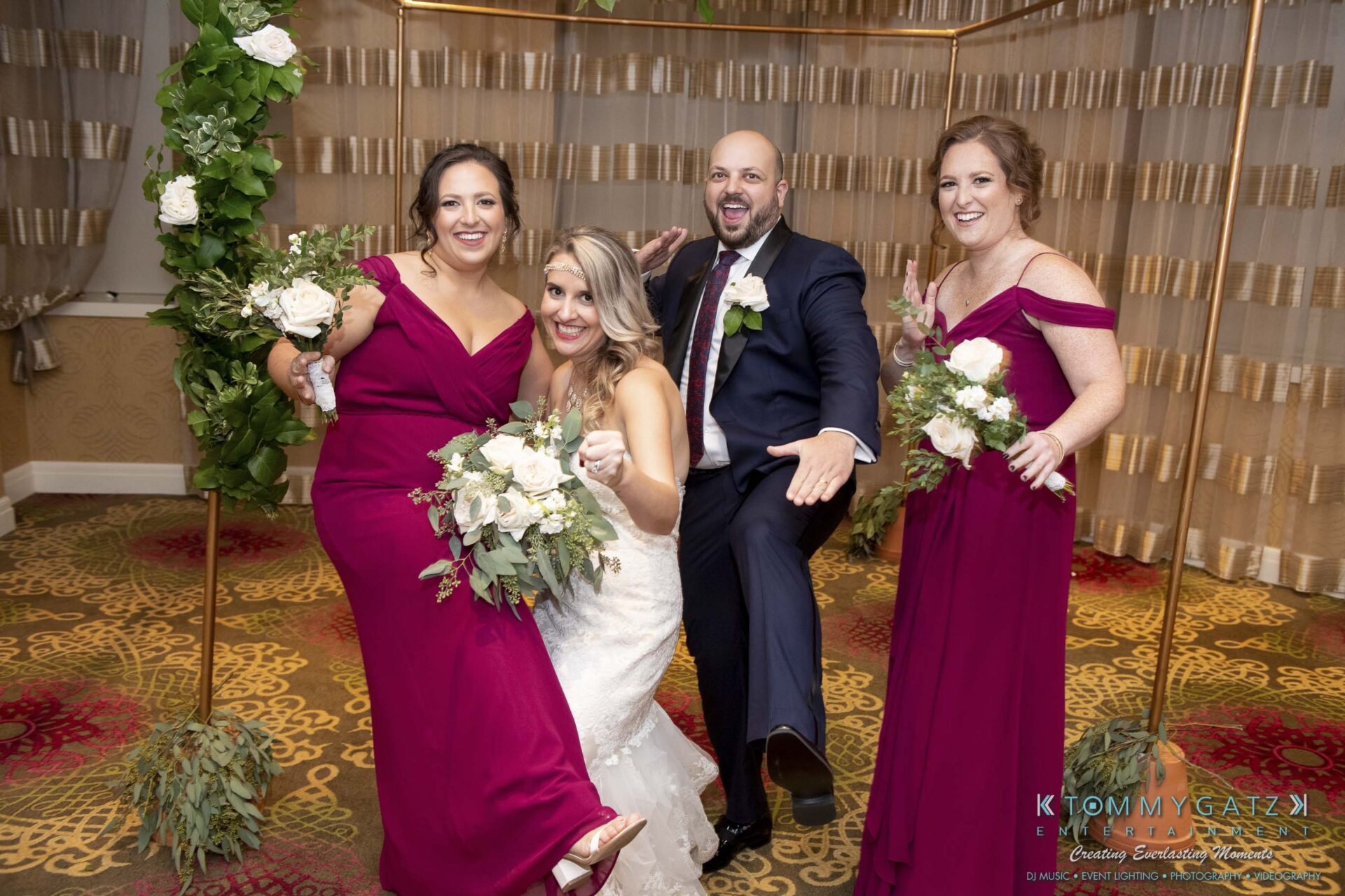 A bride and groom pose playfully with two bridesmaids, all holding white floral bouquets. They are dressed in formal attire and standing indoors.