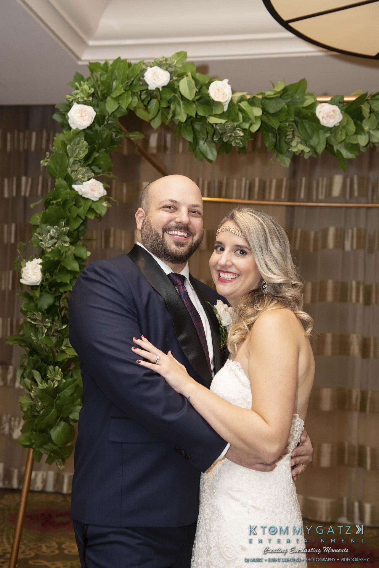 A couple in formal attire poses under a floral arch. The man wears a navy suit, and the woman wears a white dress. They smile with arms around each other.