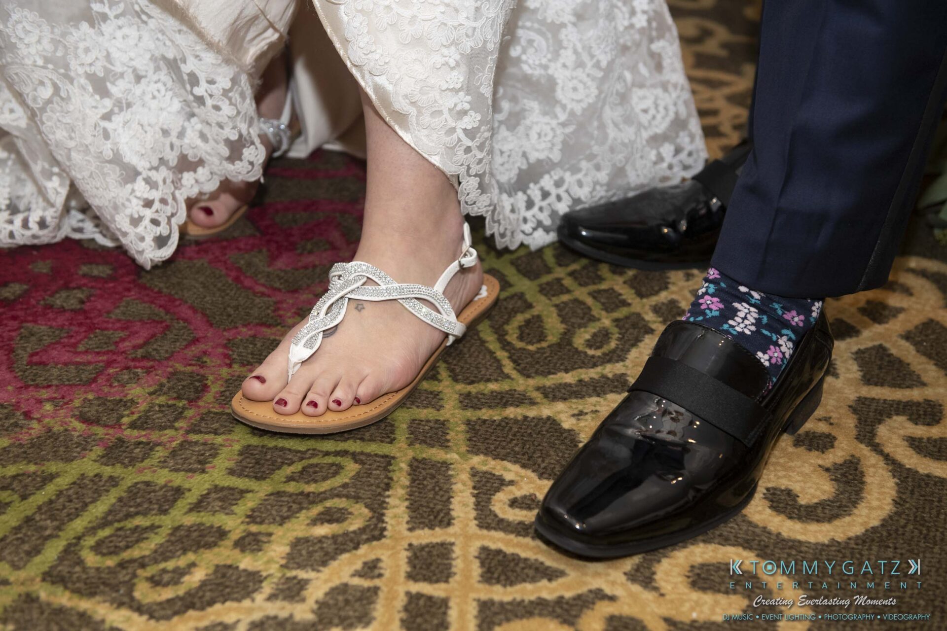 A bride's foot in a jeweled sandal next to a groom's polished shoe with floral socks on a patterned carpet.