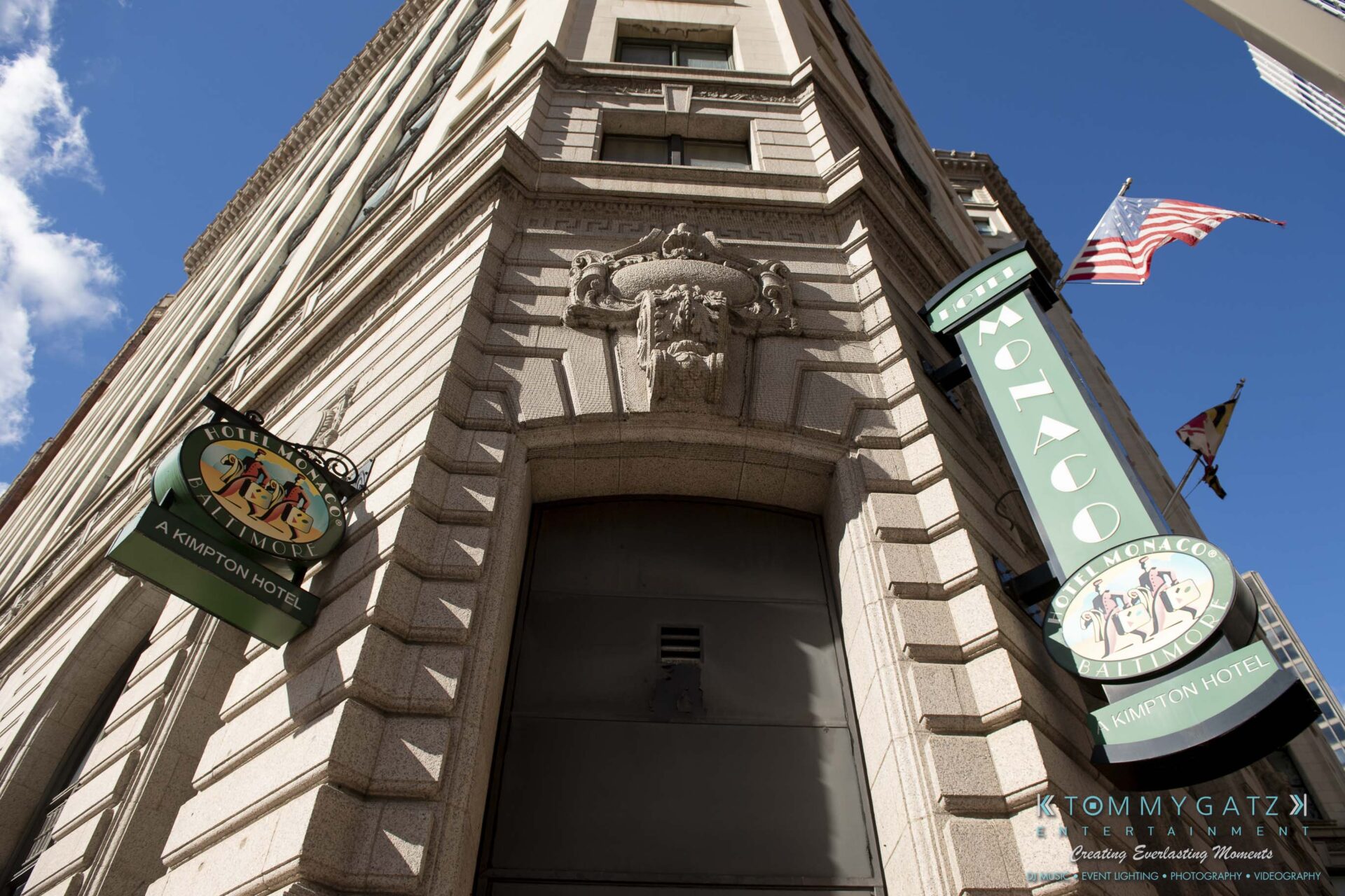 View of a tall building corner with Hotel Monaco signage. An American flag is visible above, and the sky is clear.