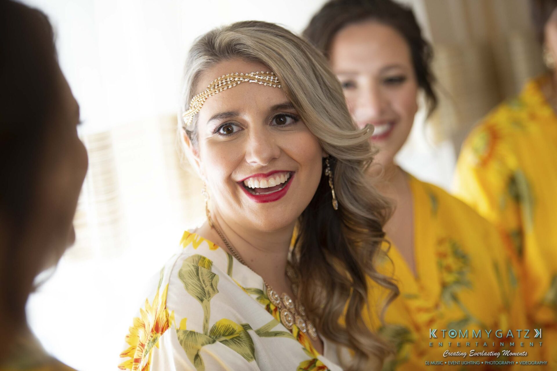 A woman with long, wavy hair and a decorative headpiece smiles while wearing a yellow floral robe. Two others in similar robes are in the background.