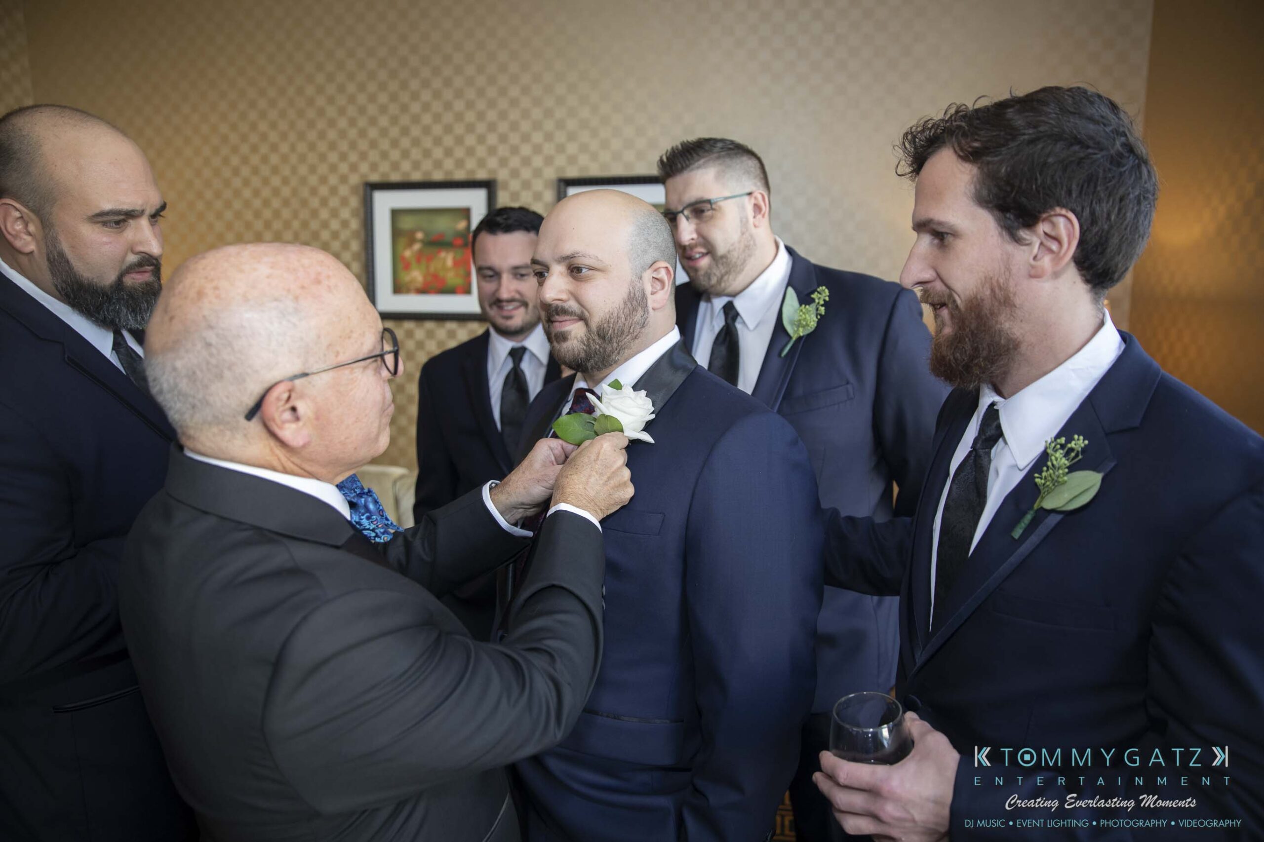 A group of formally dressed men helps a groom adjust his boutonniere in a room with a patterned wall.
