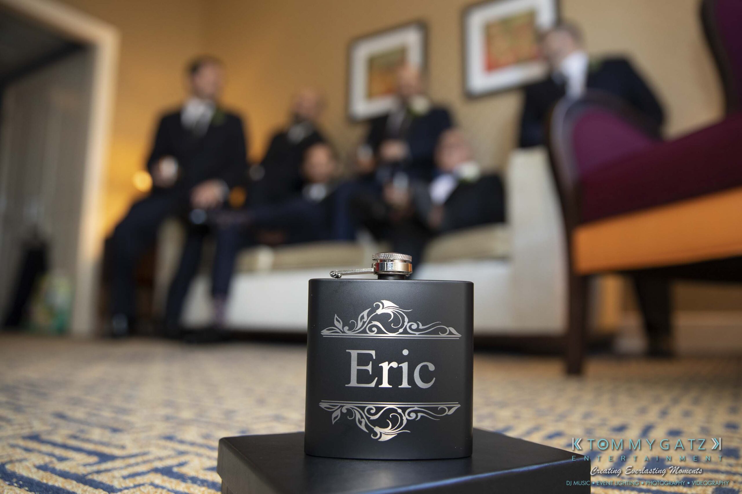 A personalized flask labeled "Eric" is in focus in the foreground, with a group of people in formal attire seated on a sofa in the blurred background.