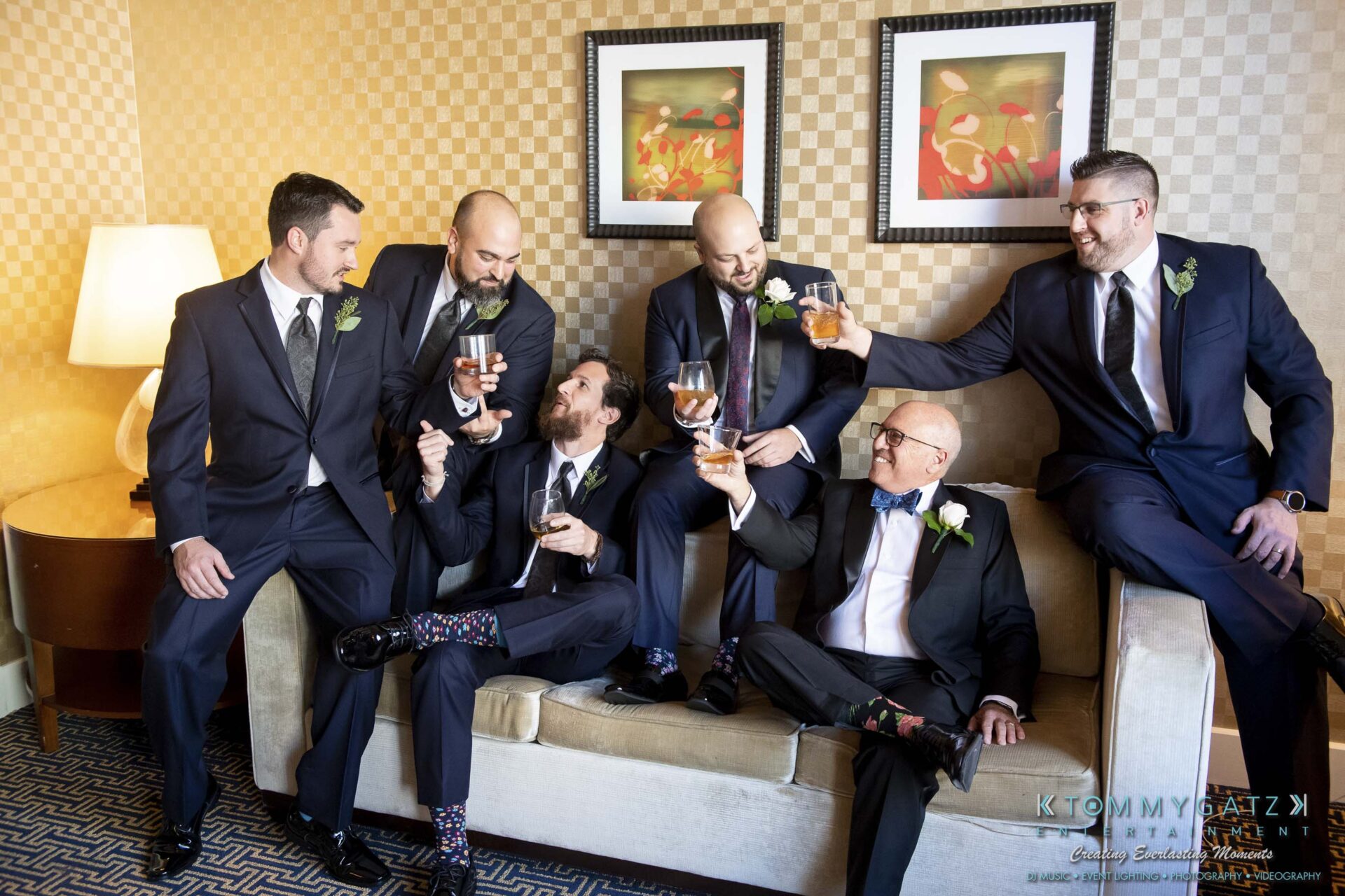 A group of six men in suits sit and stand around a sofa, holding drinks and laughing, with artwork on the wall behind them.