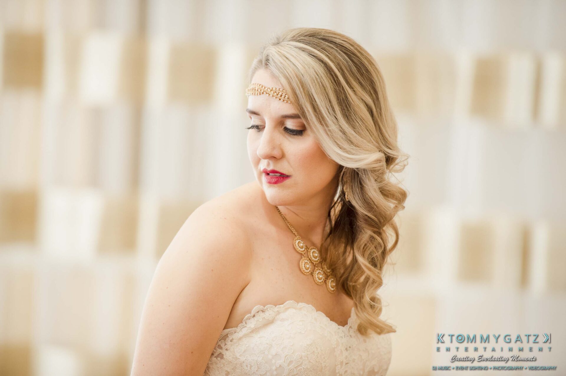 Bride in a white lace gown and gold necklace, with long blonde hair styled in waves, poses indoors looking to the side.