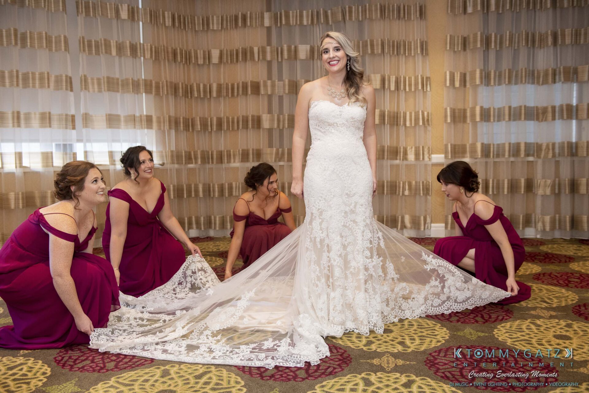 A bride in a strapless lace gown stands smiling while four bridesmaids in purple dresses adjust her train in a decorated room.