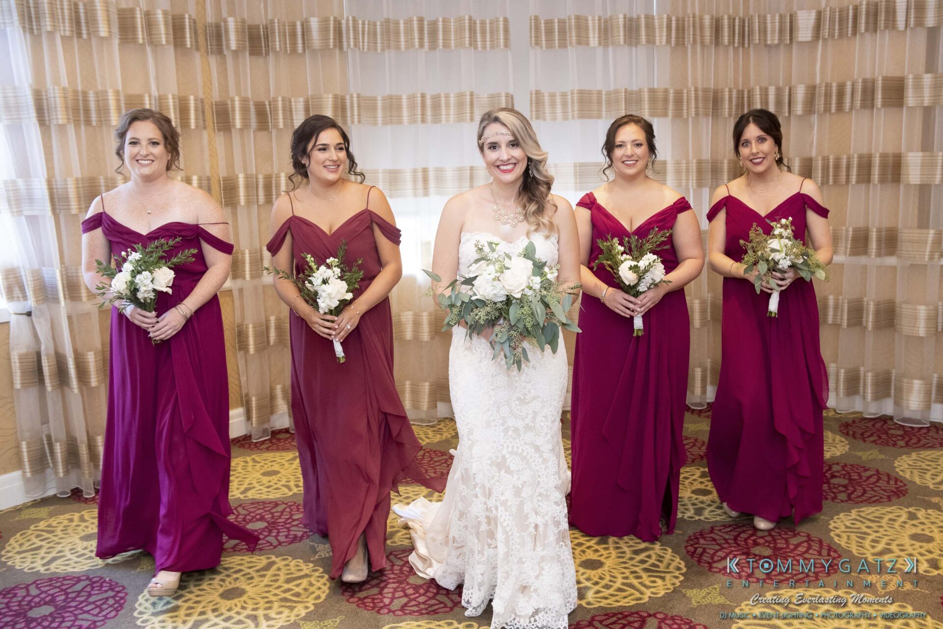 A bride in a white gown holds a bouquet, surrounded by four bridesmaids in matching burgundy dresses, each holding a bouquet, in a room with patterned carpeting and beige curtains.