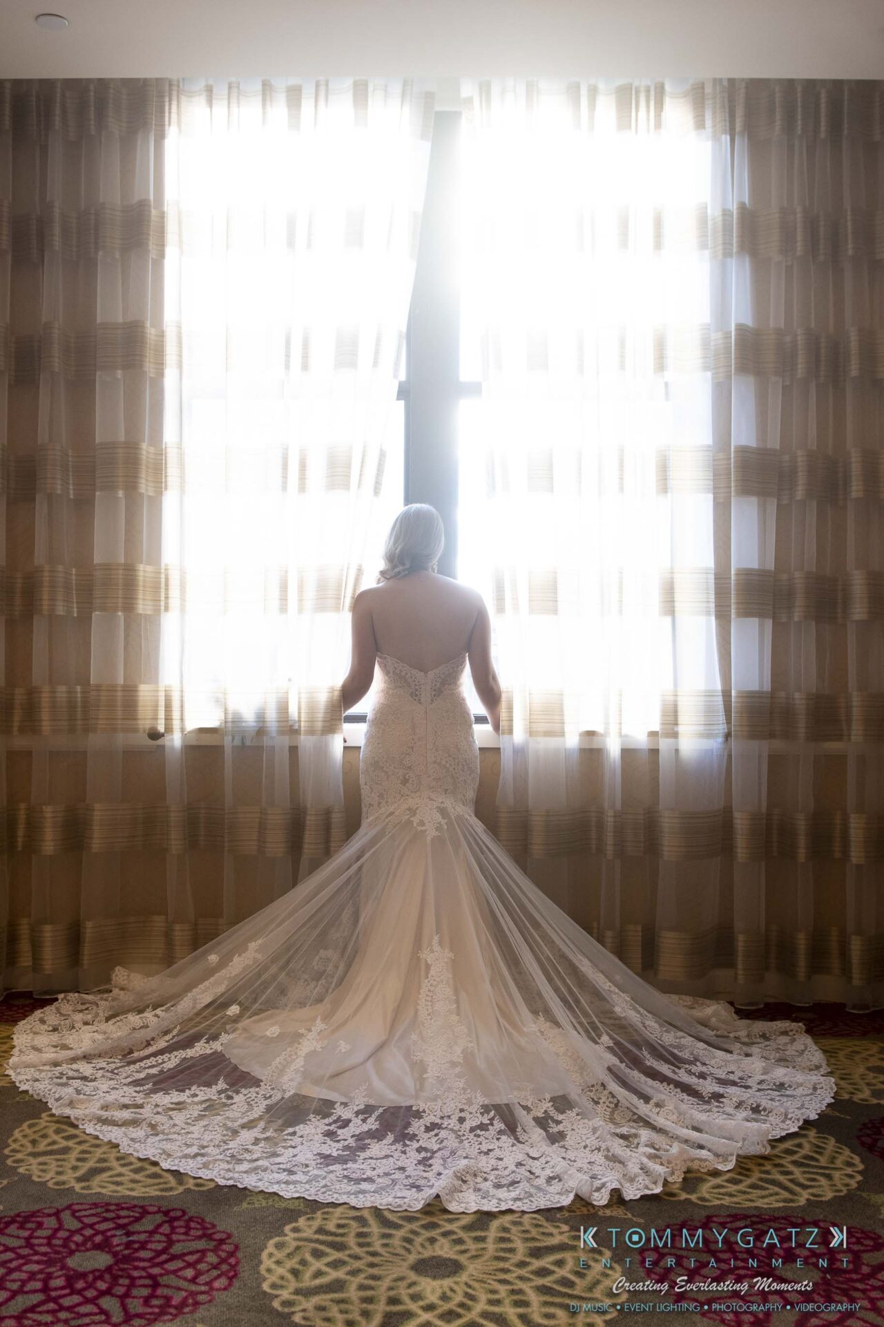 A bride in a white lace wedding gown stands facing a window, with light streaming through curtains.