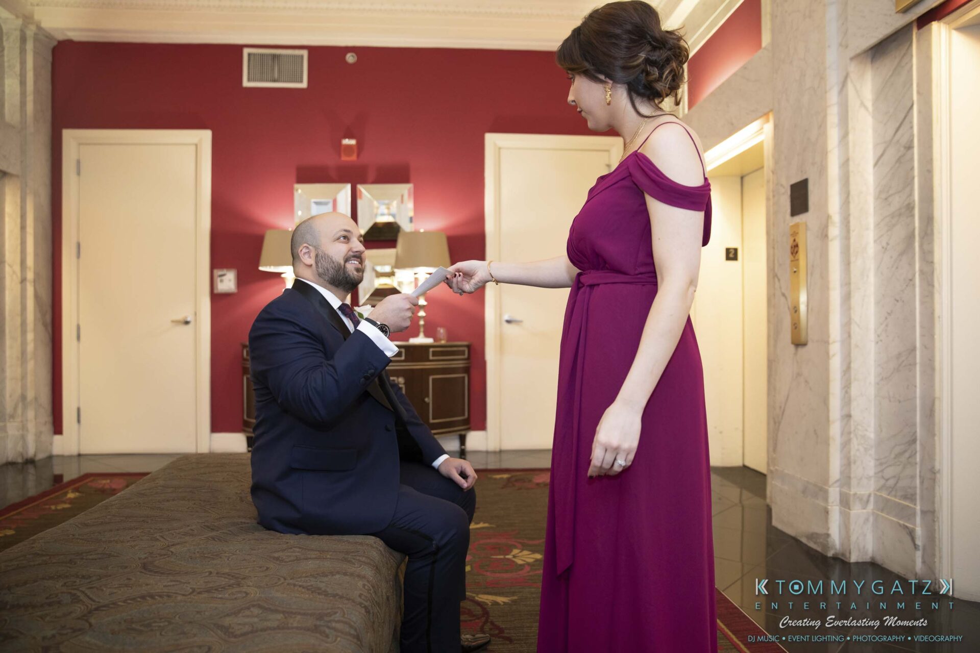 A seated man in a suit holds the hand of a standing woman in a purple dress in an elegant room with red walls.