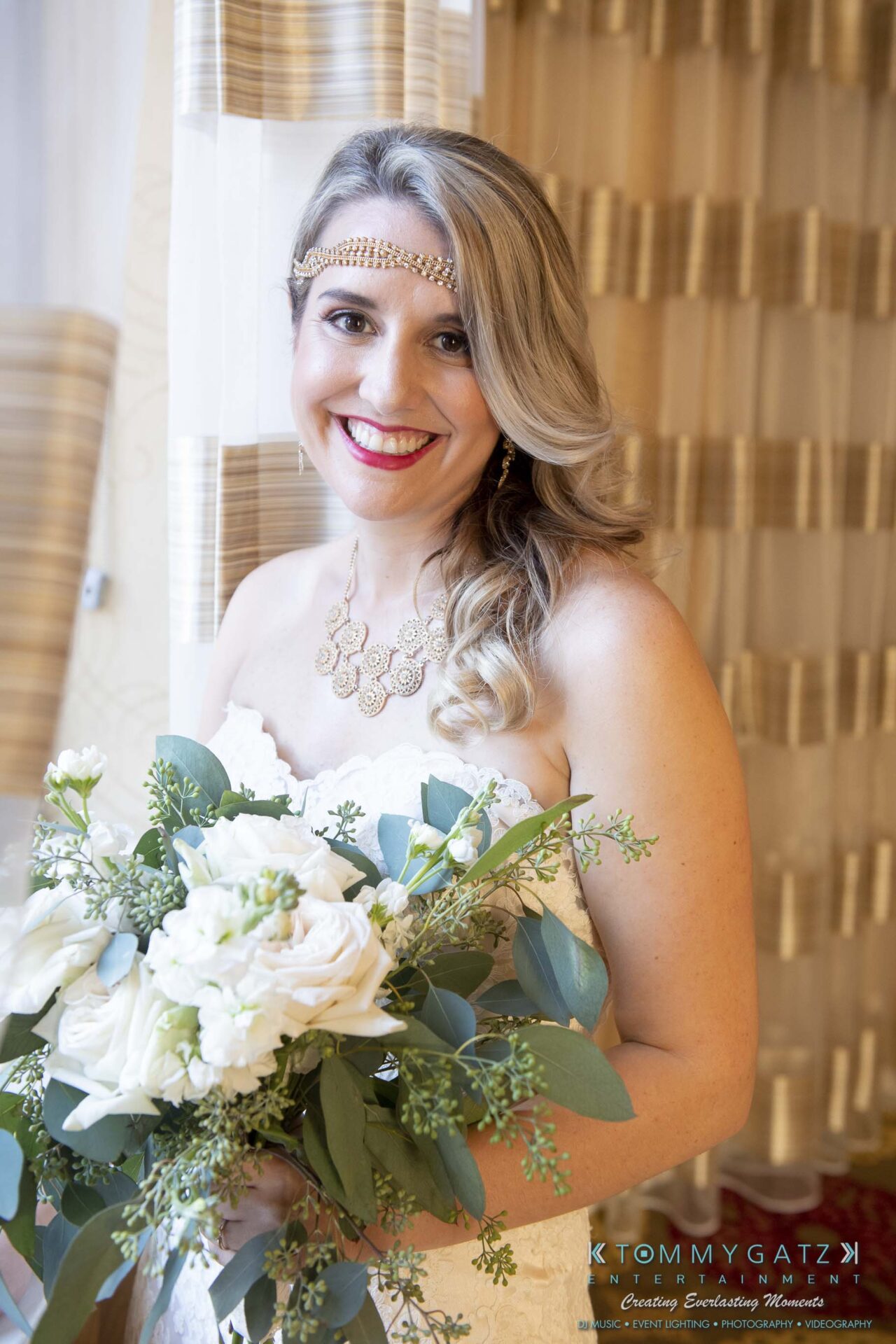 A smiling bride holding a bouquet of white flowers and greenery, wearing a gold headpiece and necklace.