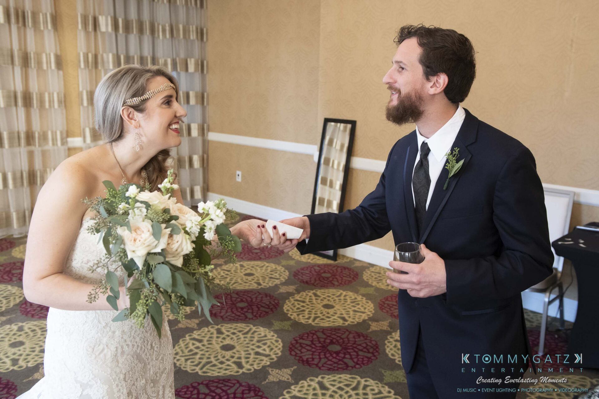 Bride and groom smiling at each other; she holds a bouquet, he holds a drink and paper. They are indoors with patterned carpet and neutral walls.