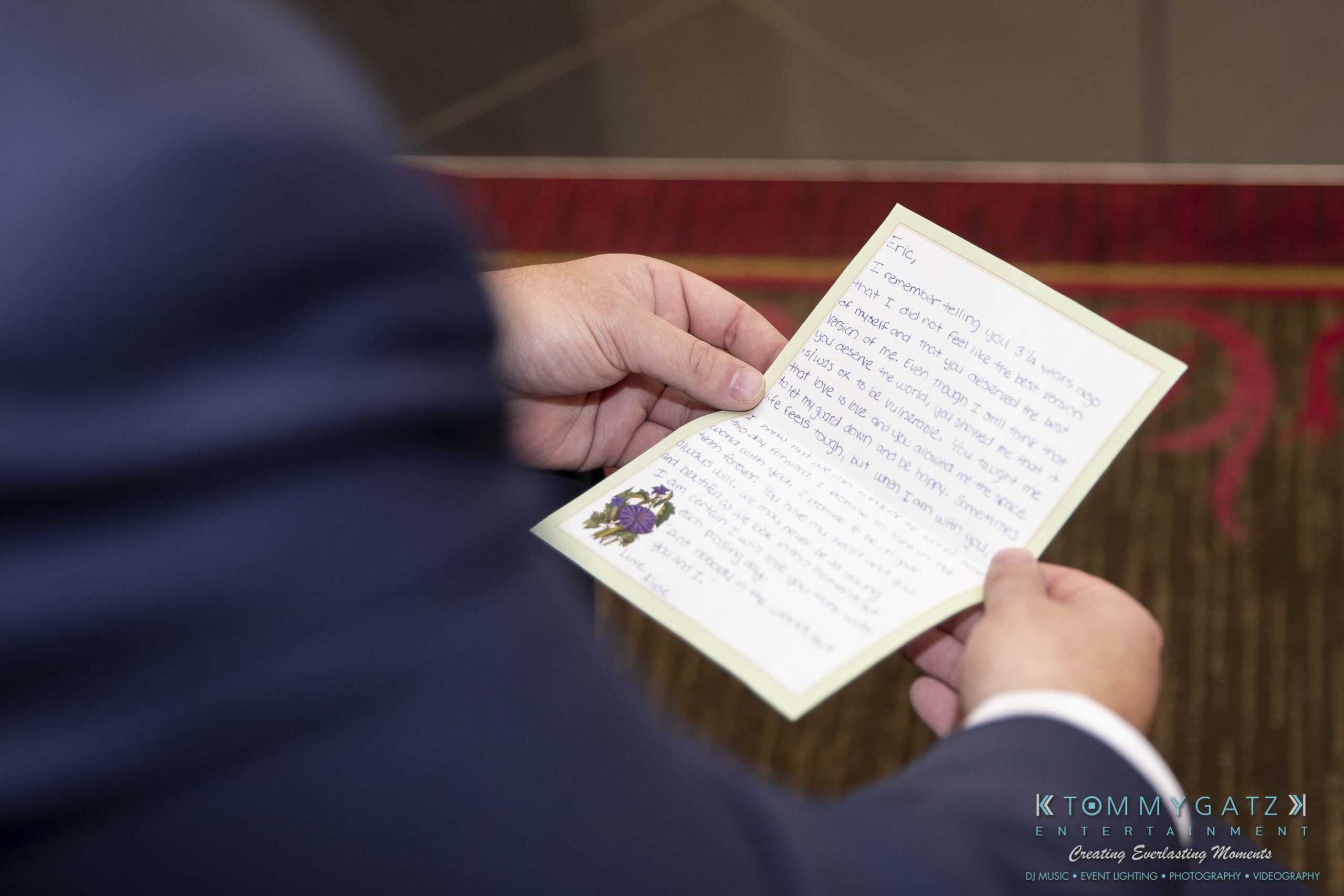 A person in a suit holds an open handwritten note decorated with a small floral design.
