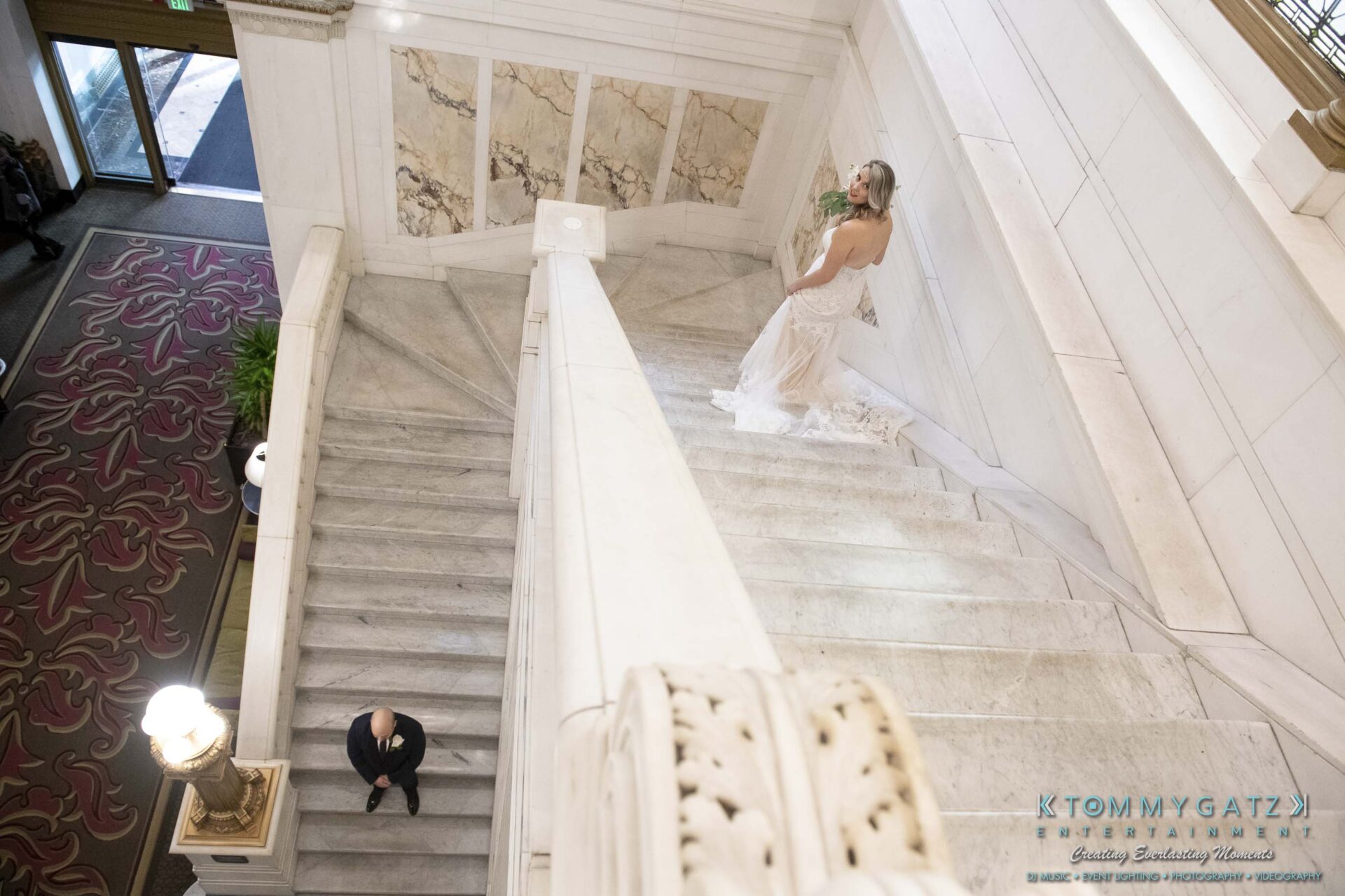 Bride in a white gown stands on a marble staircase, while a man in formal attire walks up the stairs below in an elegant building interior.