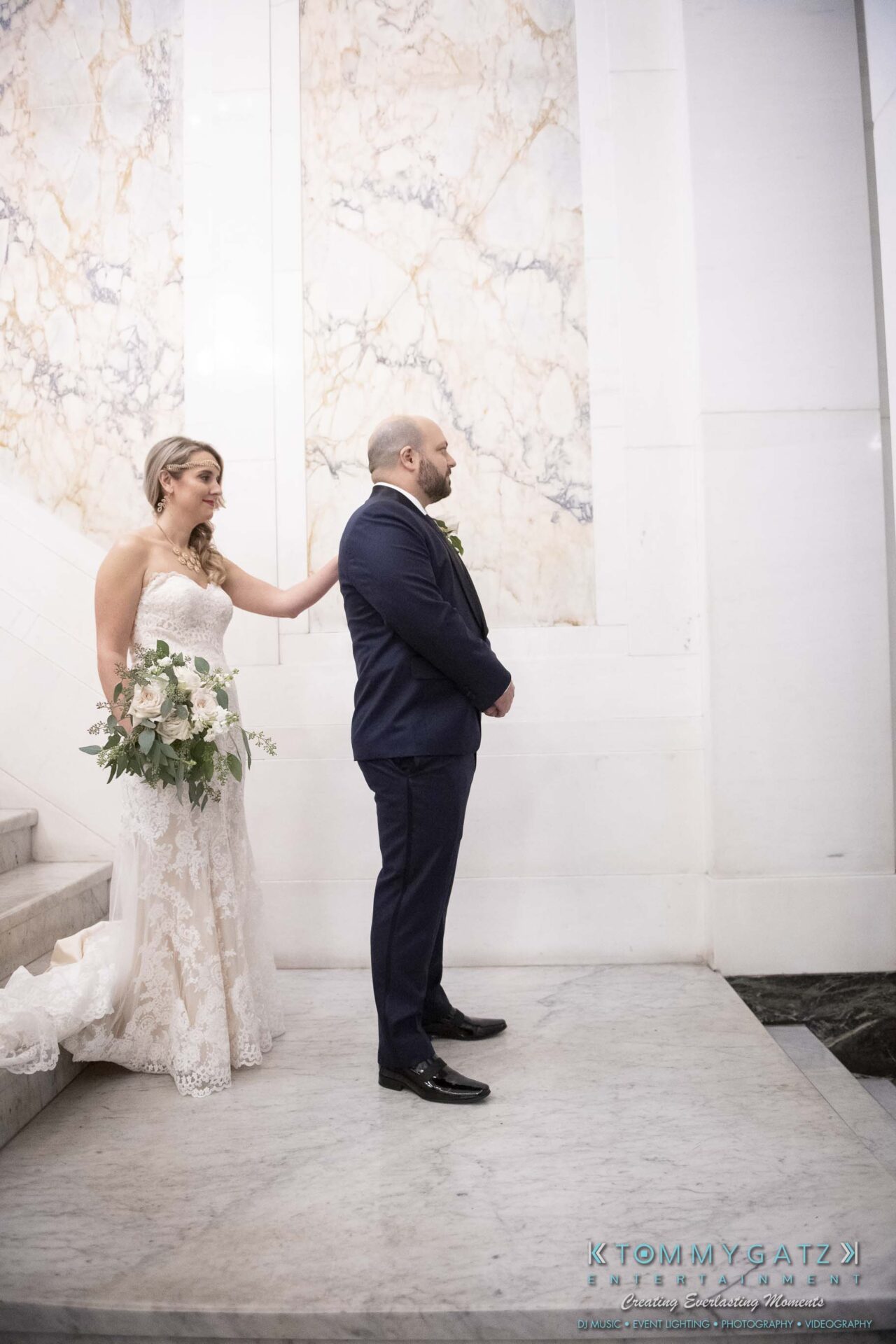 A bride in a lace gown gently taps the groom on the shoulder. They stand on a marble staircase, preparing for a first look.
