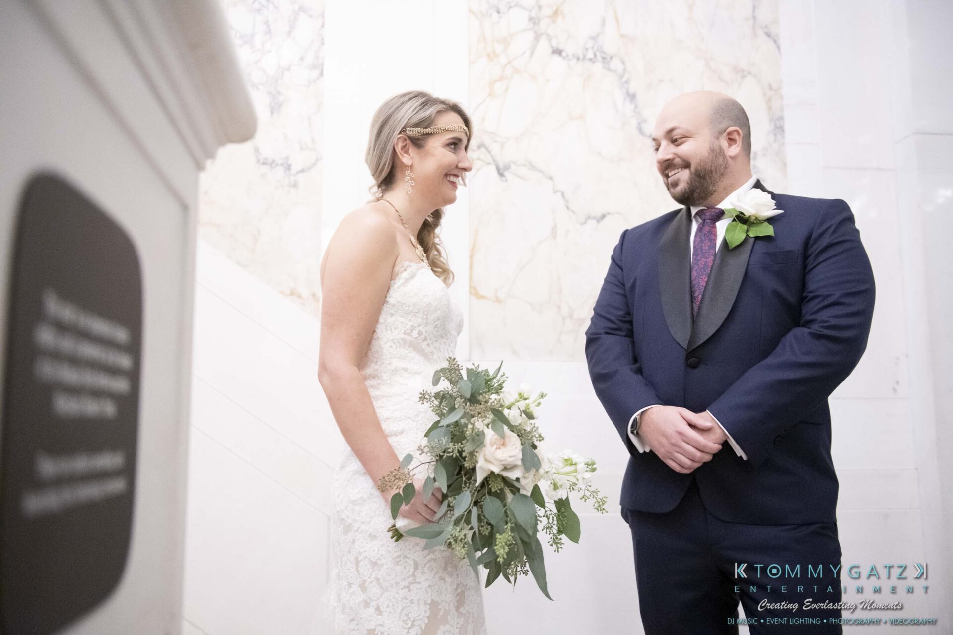 A bride and groom smile at each other on a marble staircase. The bride holds a bouquet, and both are dressed in formal attire.