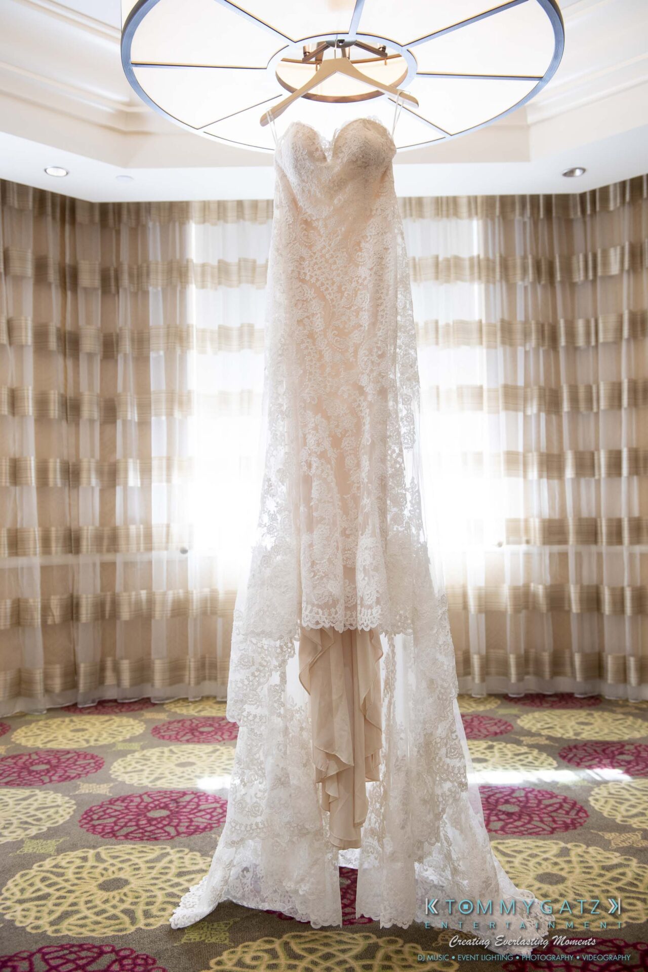 A lace wedding gown hangs from a circular ceiling light fixture in a room with patterned carpet and sheer curtains.