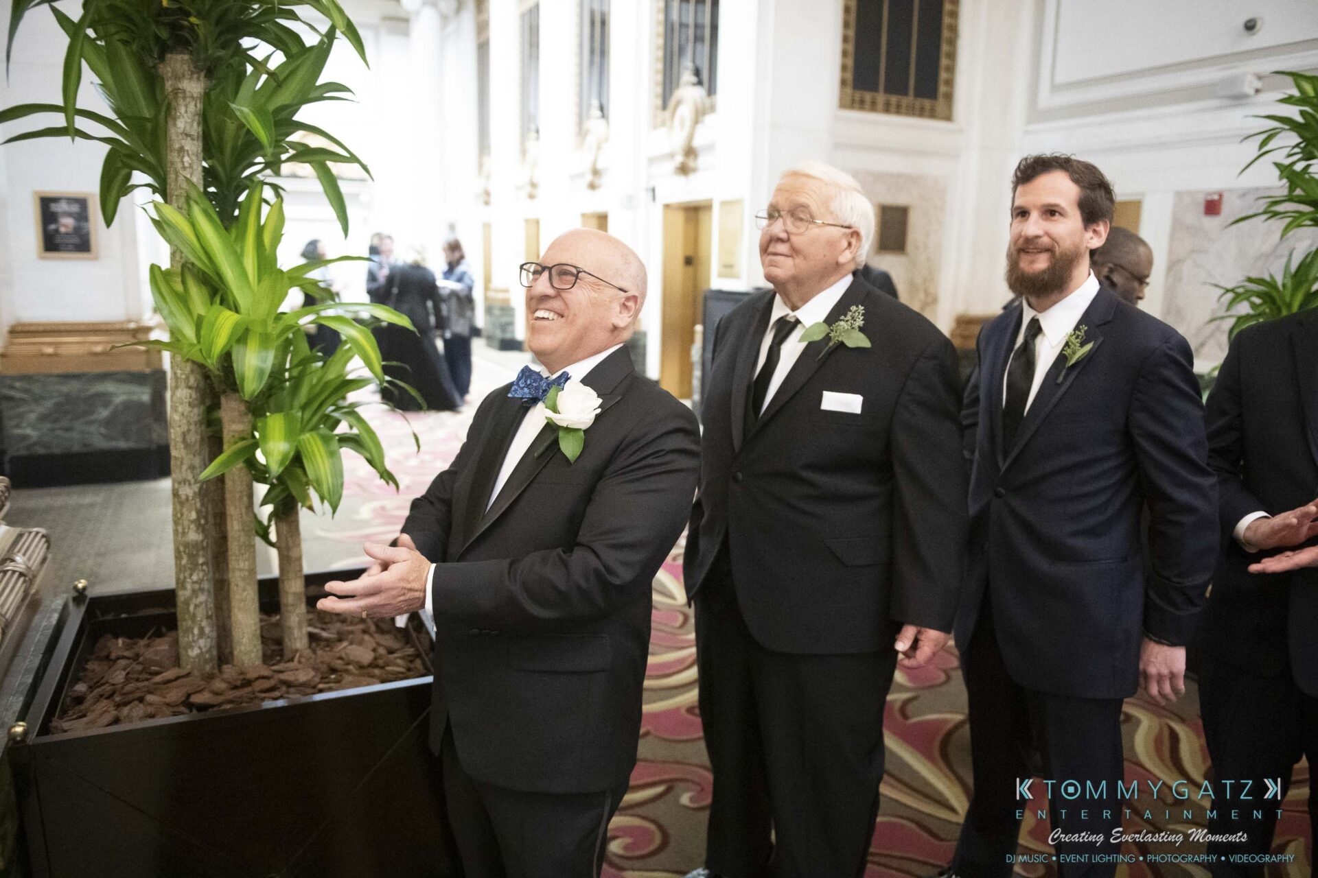Three men in suits stand in a formal setting, smiling and looking ahead. The man on the left holds a boutonniere.