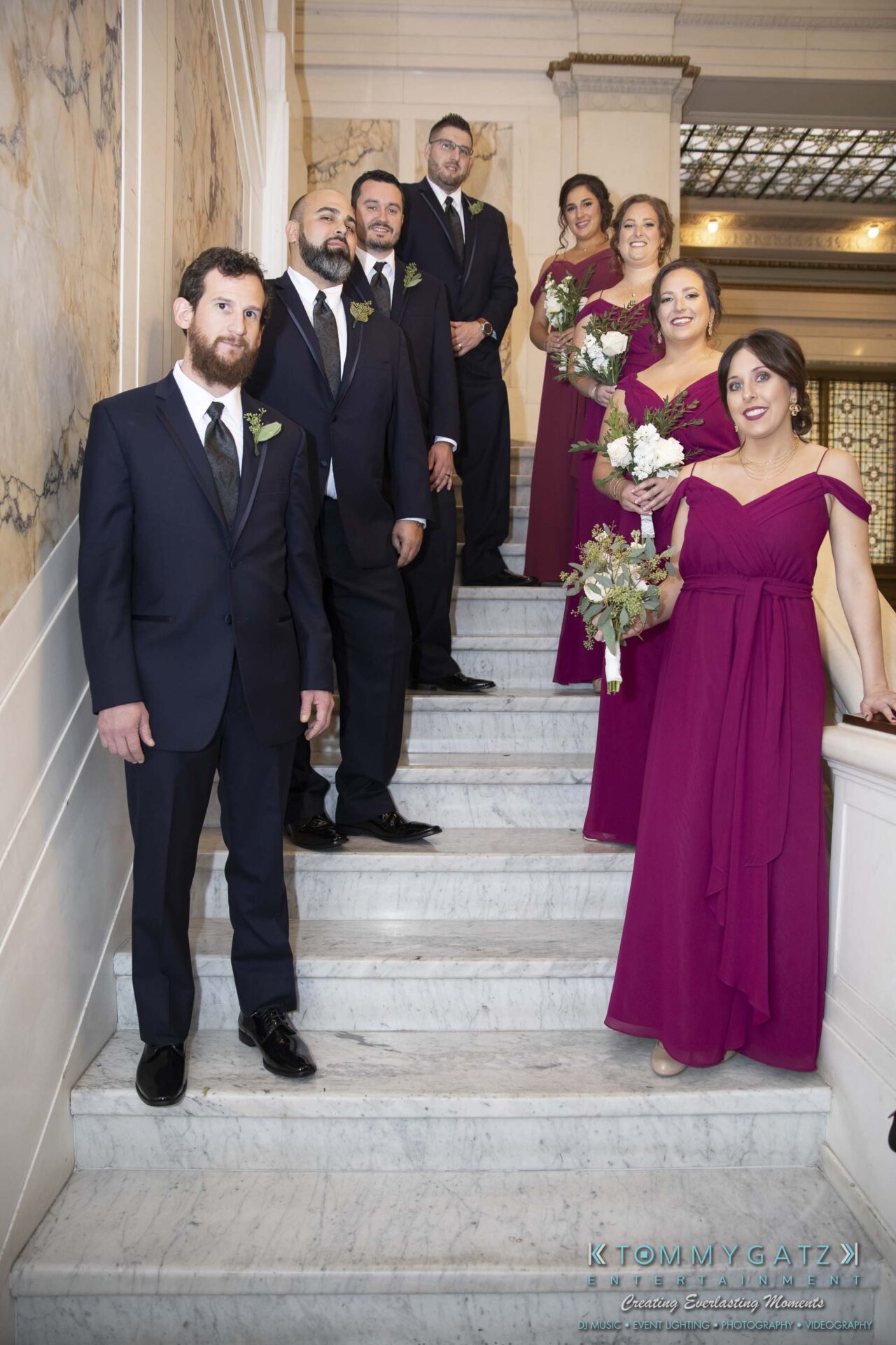 A group of people, with men in suits and women in purple dresses holding bouquets, standing on a marble staircase indoors.