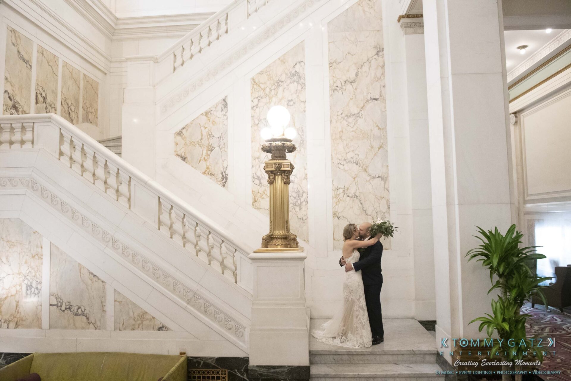 A bride and groom embrace on the steps of an ornate marble staircase, with a vintage lamp and decorative plants nearby.