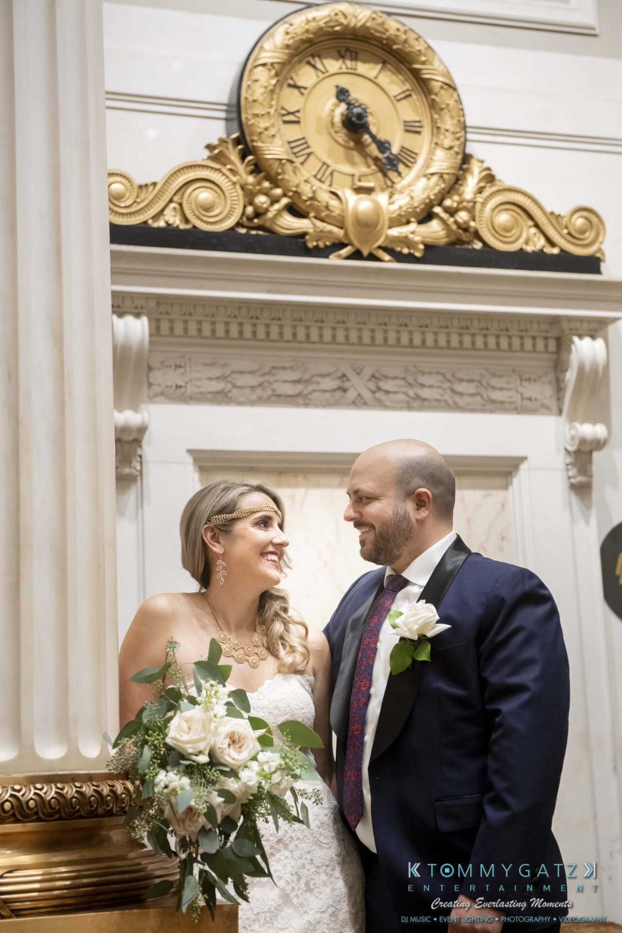 A bride and groom smile at each other in an elegant venue with a gold and black clock in the background. The bride holds a bouquet of white flowers.