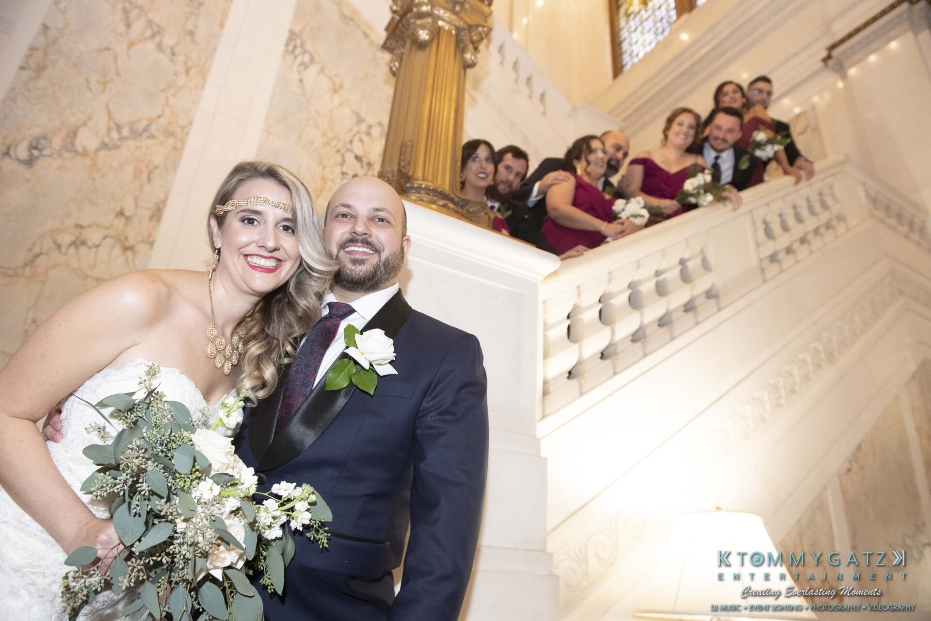 Bride and groom smiling on a staircase with a wedding party posing behind them, holding bouquets.
