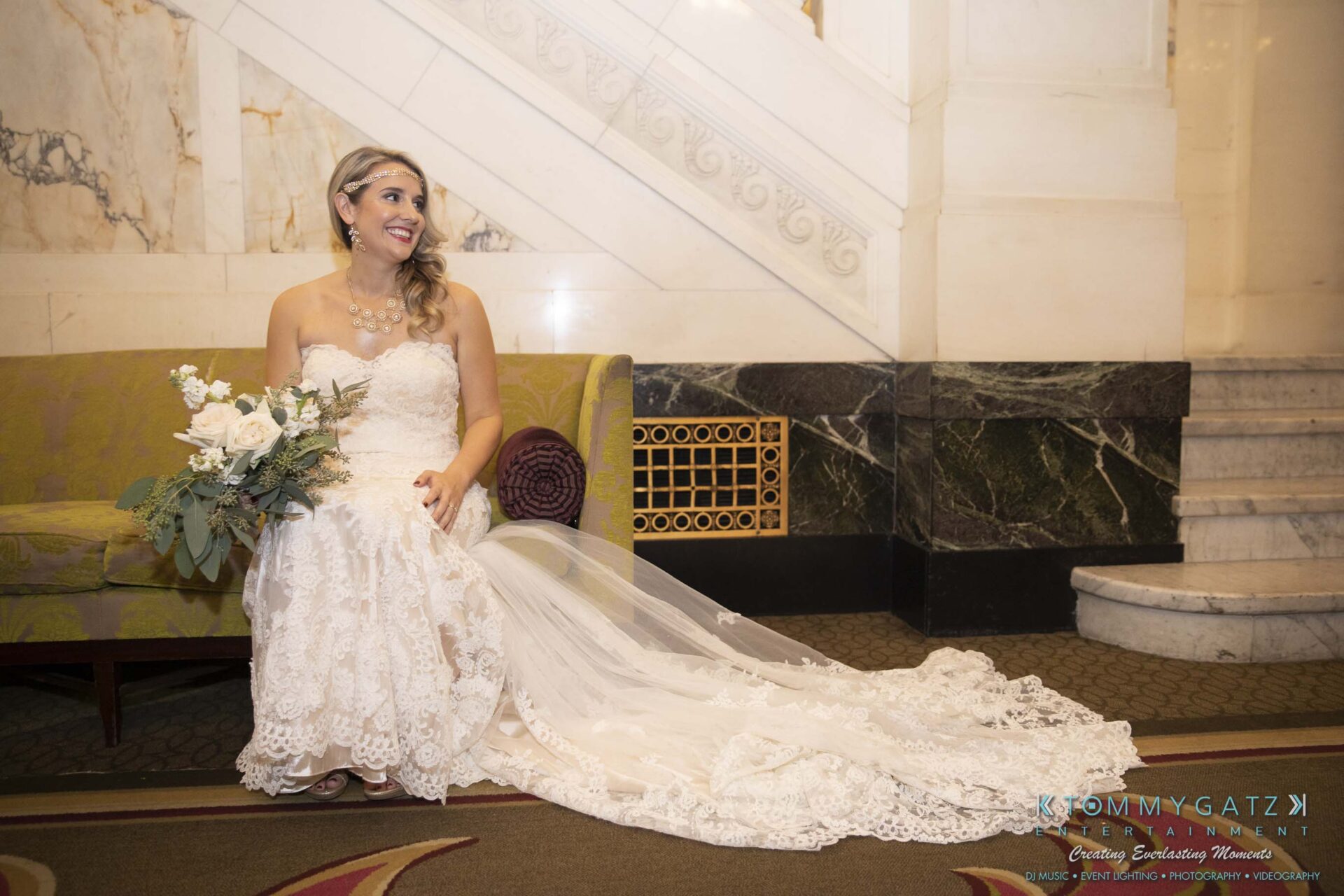Bride sitting on a couch in an ornate room, wearing a white lace wedding dress with a long train, holding a bouquet of white roses and greenery, and smiling.