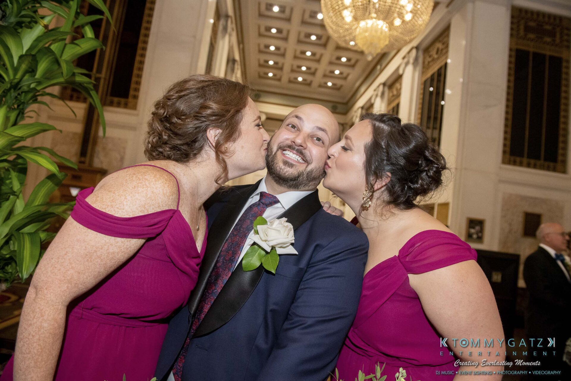 Two women in matching magenta dresses kiss a smiling man in a suit and tie on the cheeks, in an elegant venue with a chandelier in the background.