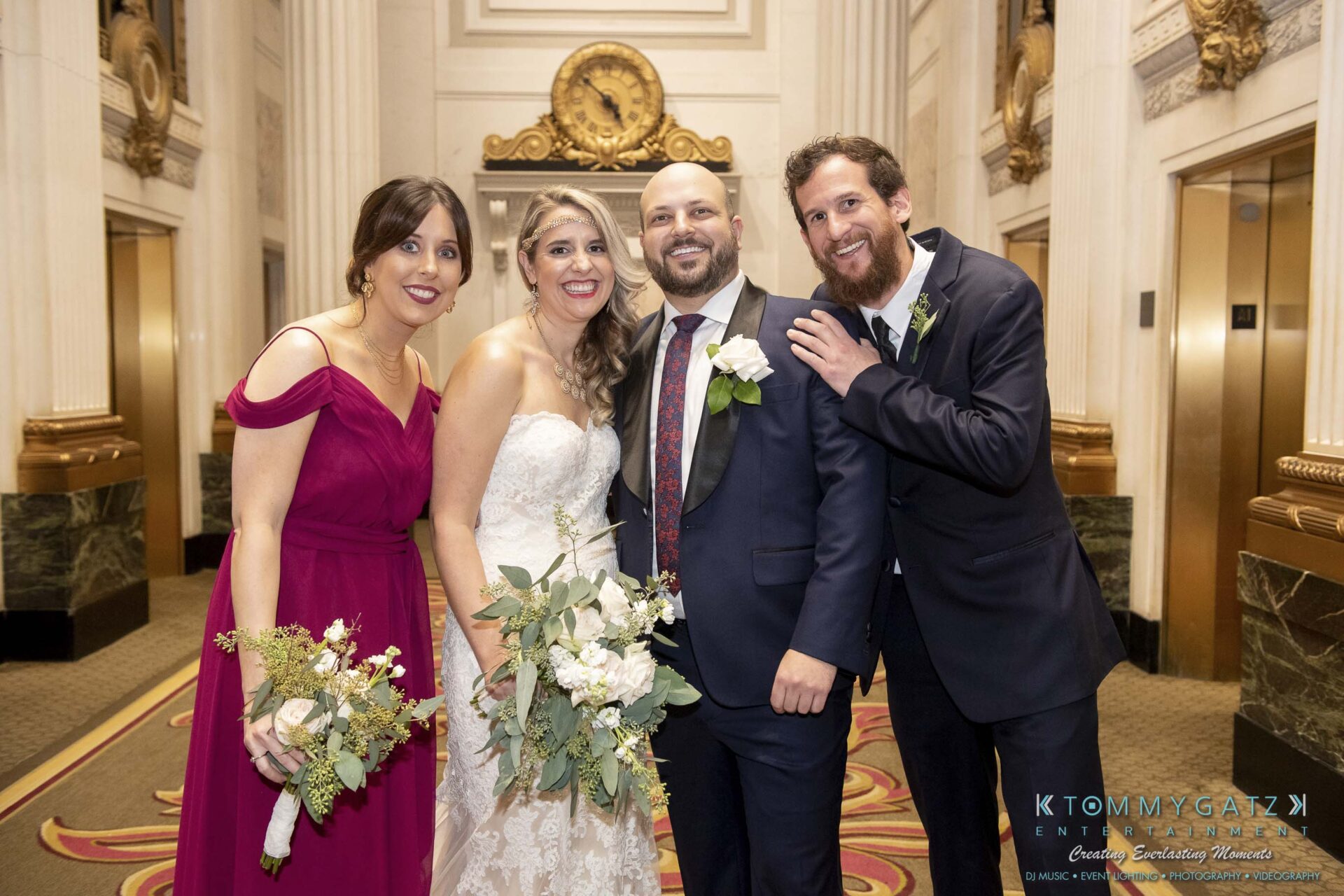 Four people smiling at a wedding, with two holding bouquets. They stand in a decorated indoor setting with large columns and ornate details.