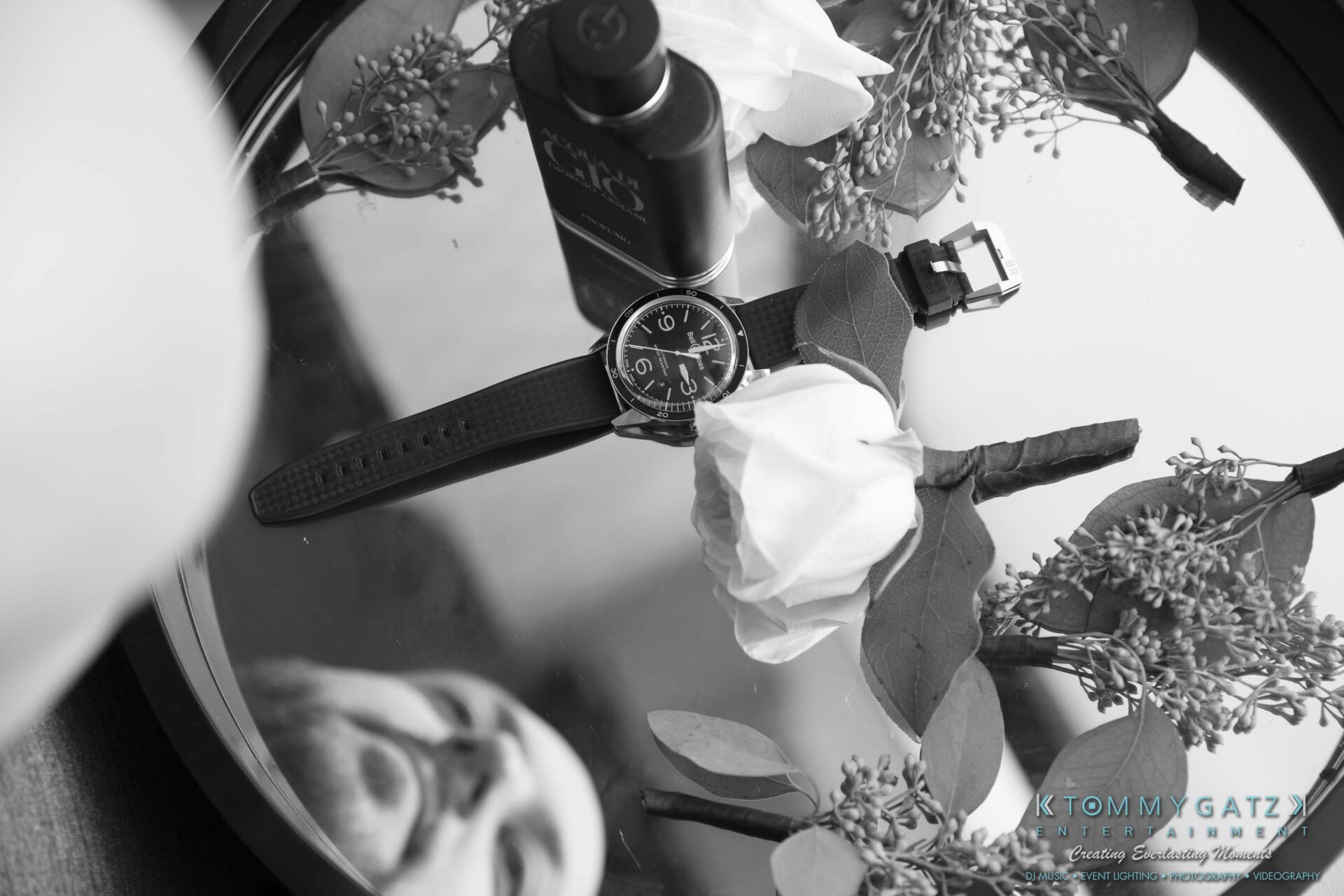 Black and white image of a wristwatch, perfume bottle, and flowers arranged on a reflective surface, with a partial reflection of a person's face visible.