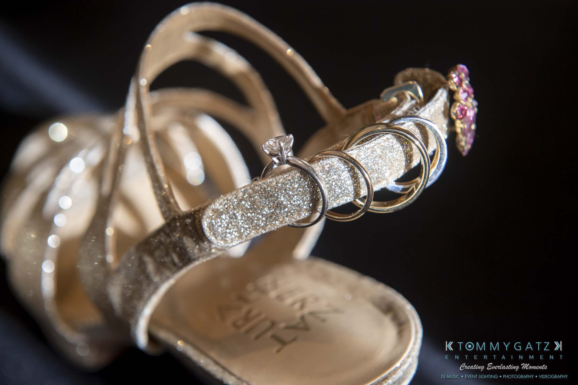 Close-up of a sparkling high-heeled sandal adorned with multiple rings, including one with a diamond, against a dark background.