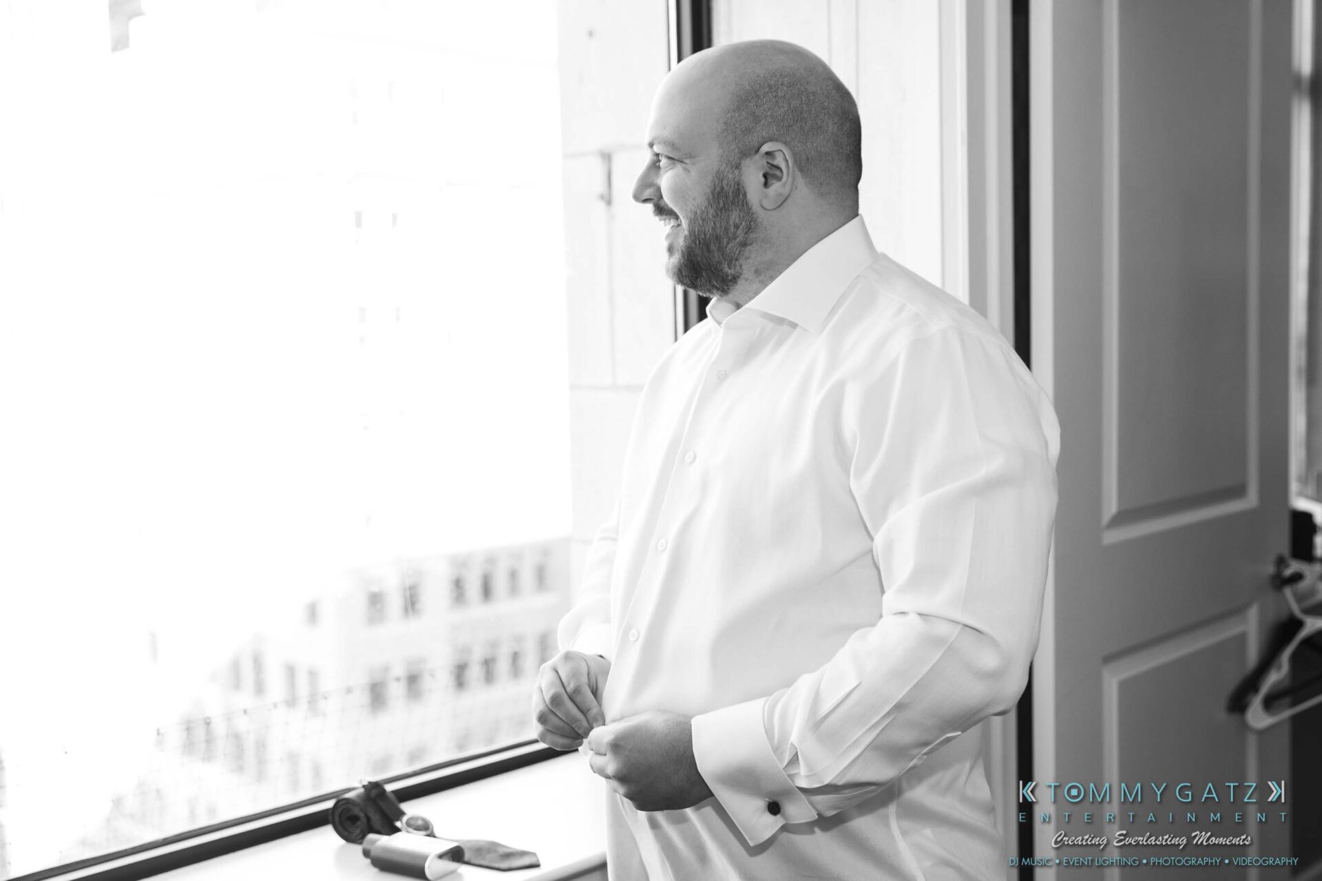 A man in a white shirt stands by a window, looking outside with a slight smile.