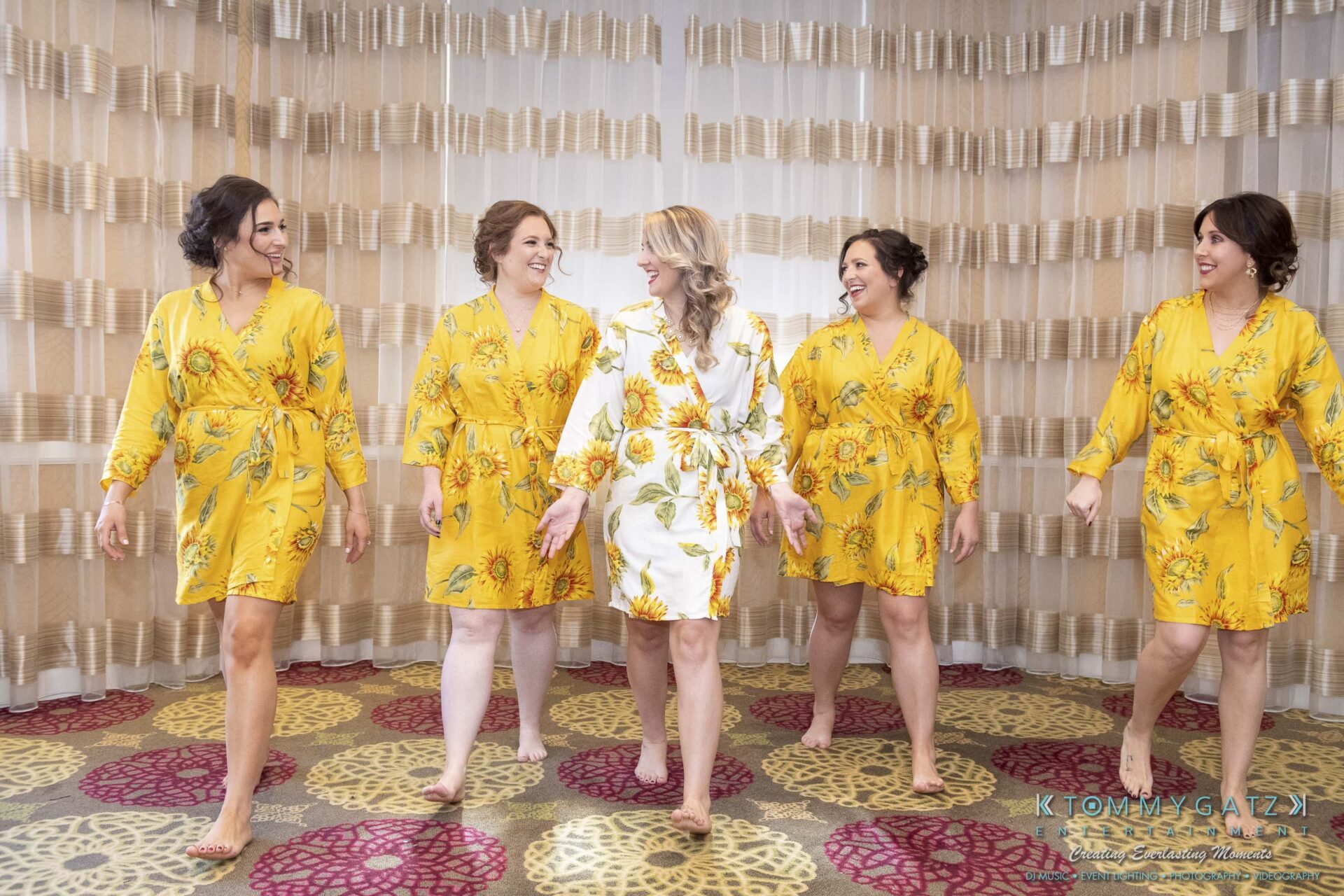 Five women in floral robes smile and walk in a room with patterned curtains and carpet. Four wear yellow robes, and one wears a white robe.