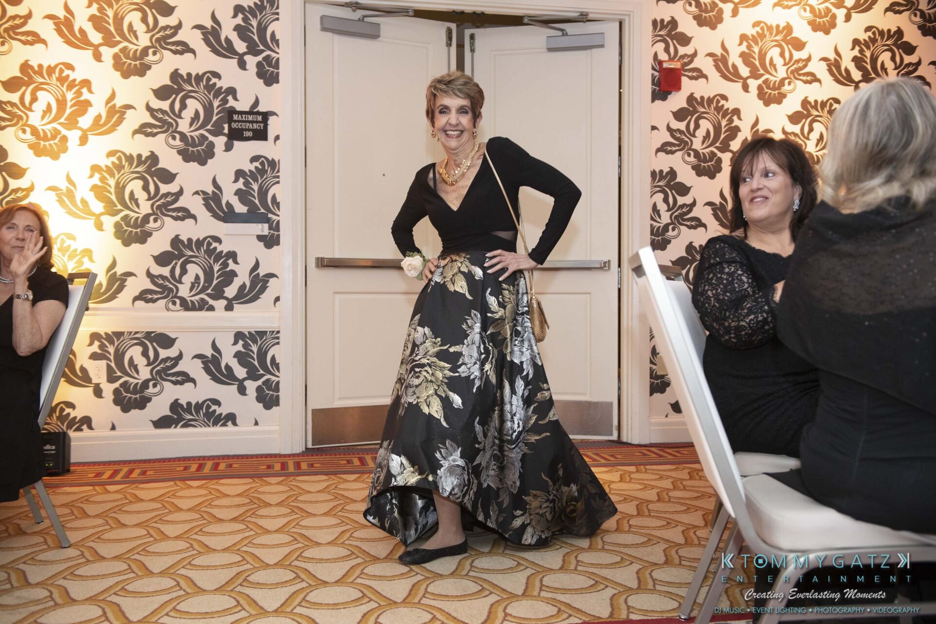 A woman poses confidently in an elegant floral skirt and black top at a formal event, surrounded by seated attendees in a ballroom.