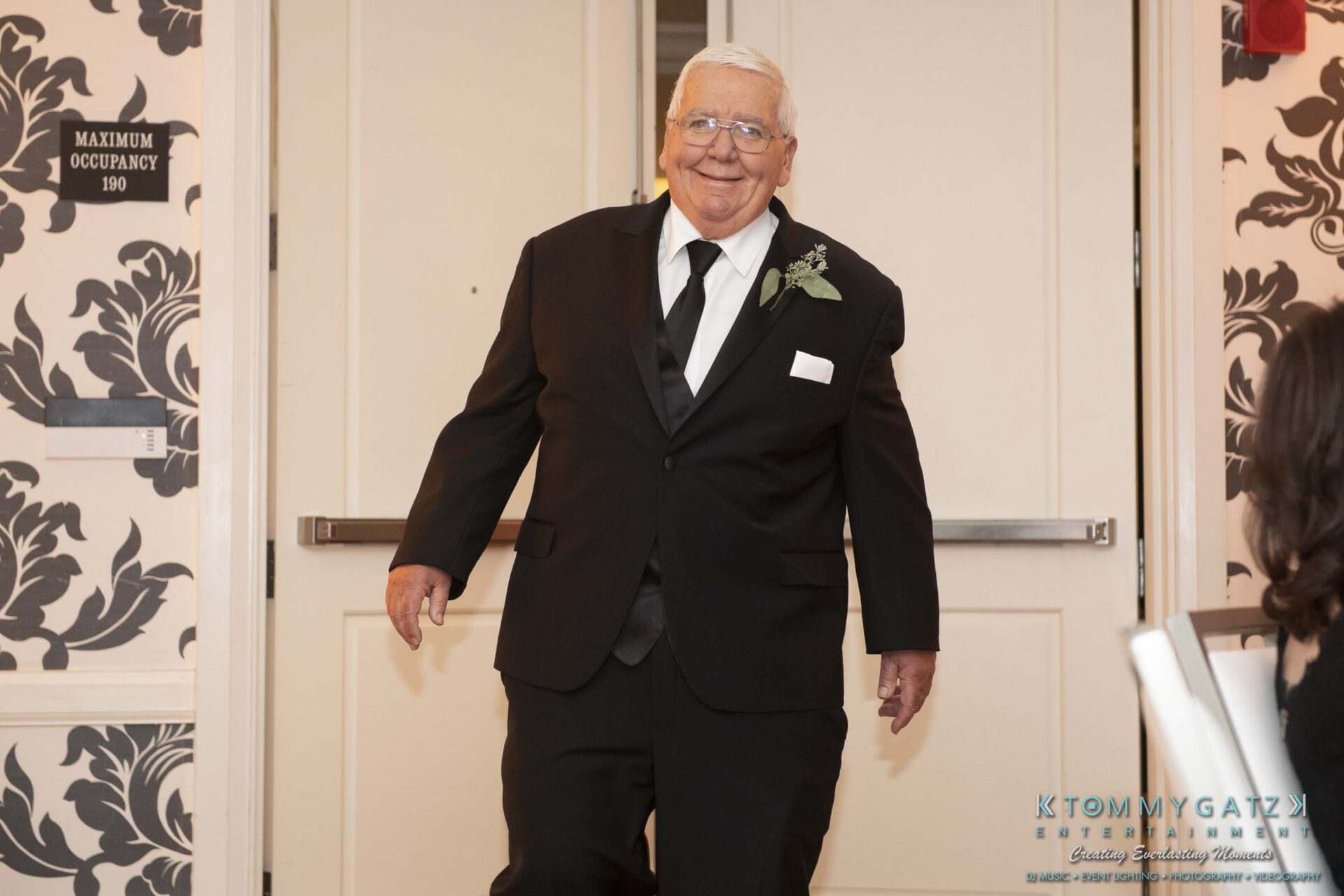 An elderly man in a black suit and tie enters a room, smiling. A boutonniere is pinned to his lapel.