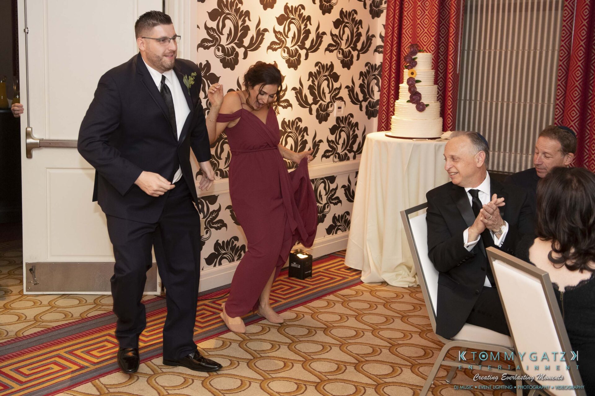 Two people in formal attire dance into a decorated room at an event, while seated guests watch. A tiered cake is visible in the background.