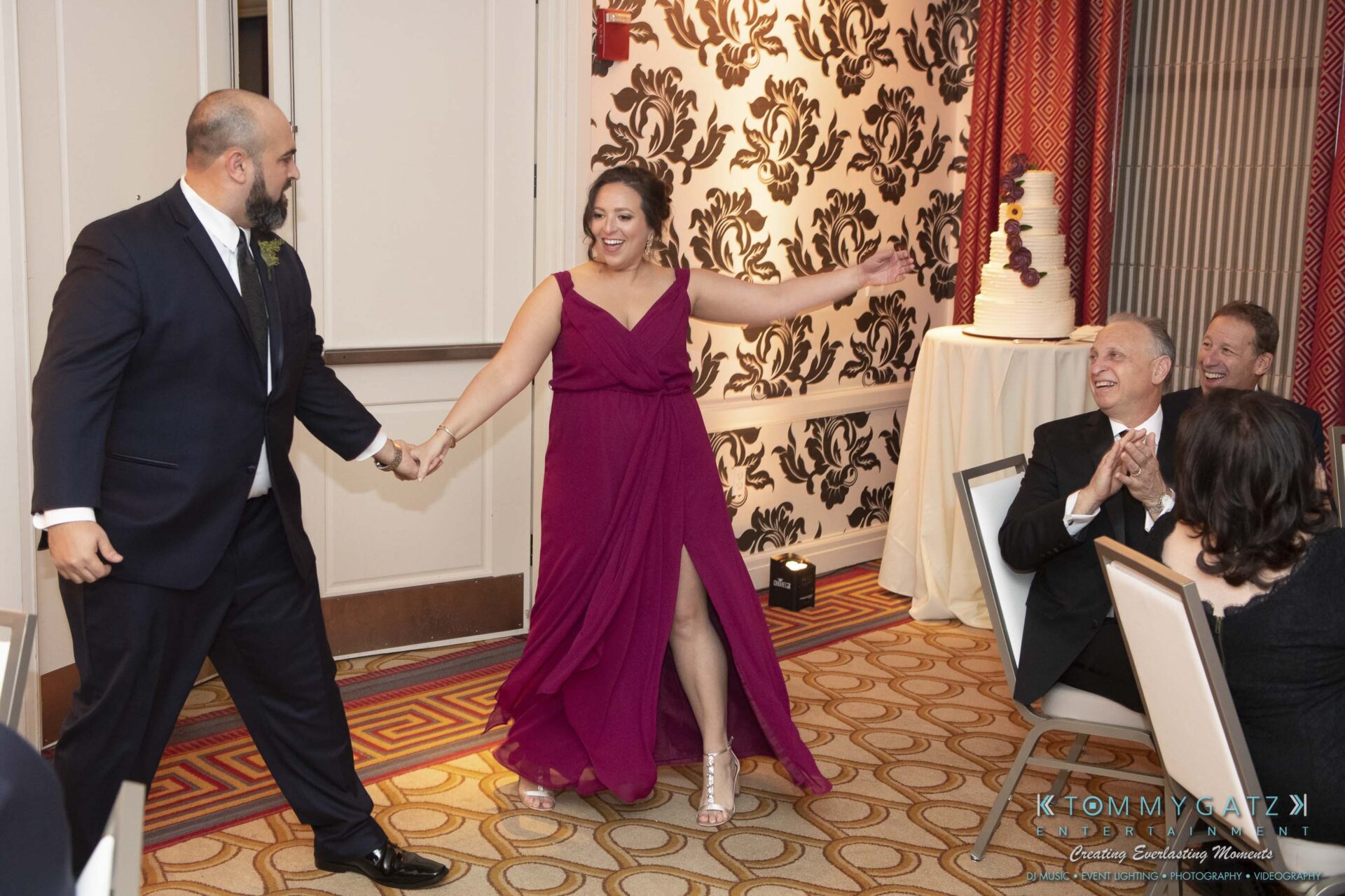 A couple holding hands enters a room; the woman wears a magenta dress and the man a dark suit. People seated nearby applaud and smile.