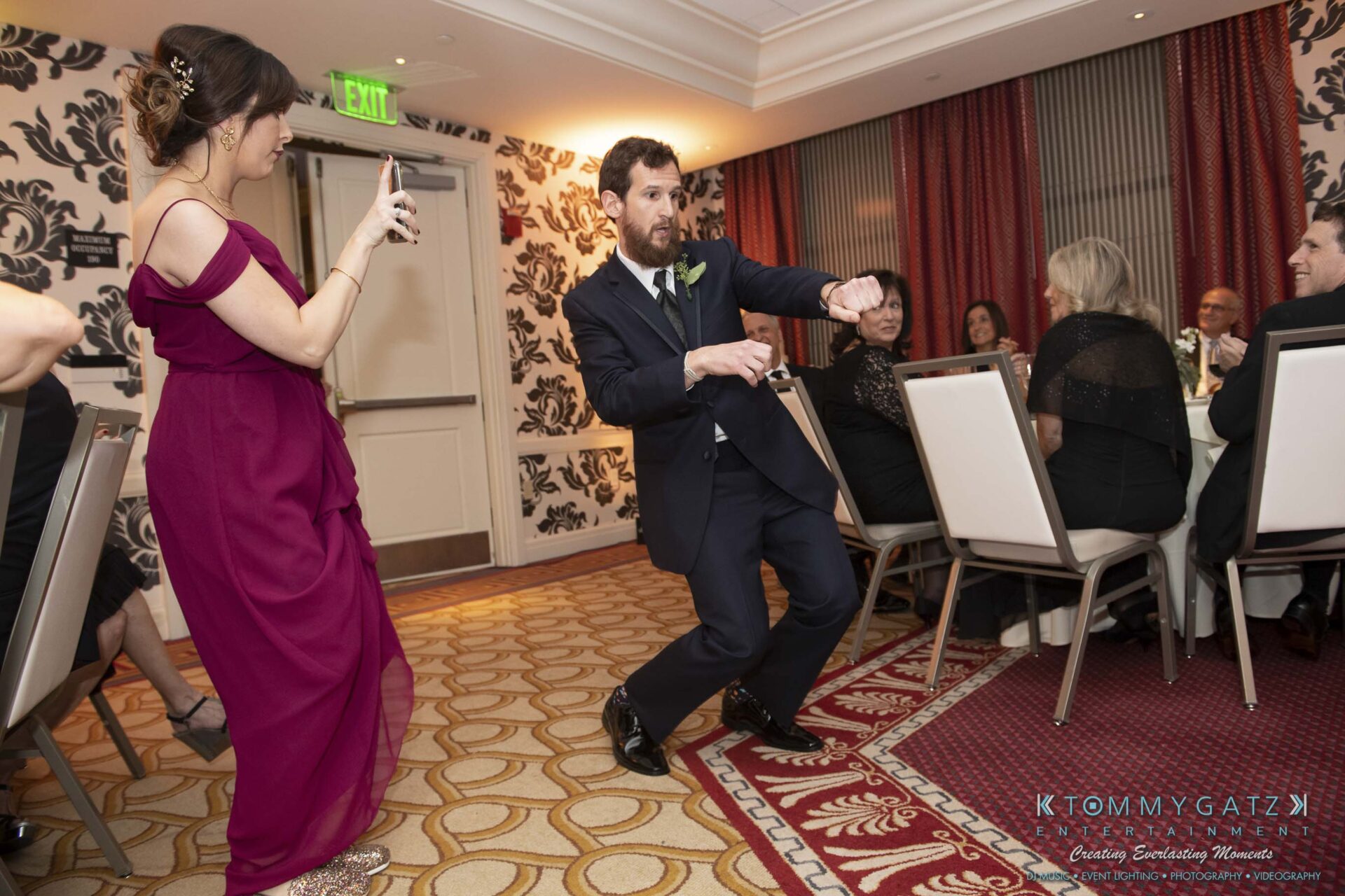 A man in a suit dances energetically in a banquet hall while a woman in a purple dress films with her phone. Guests are seated at tables in the background.