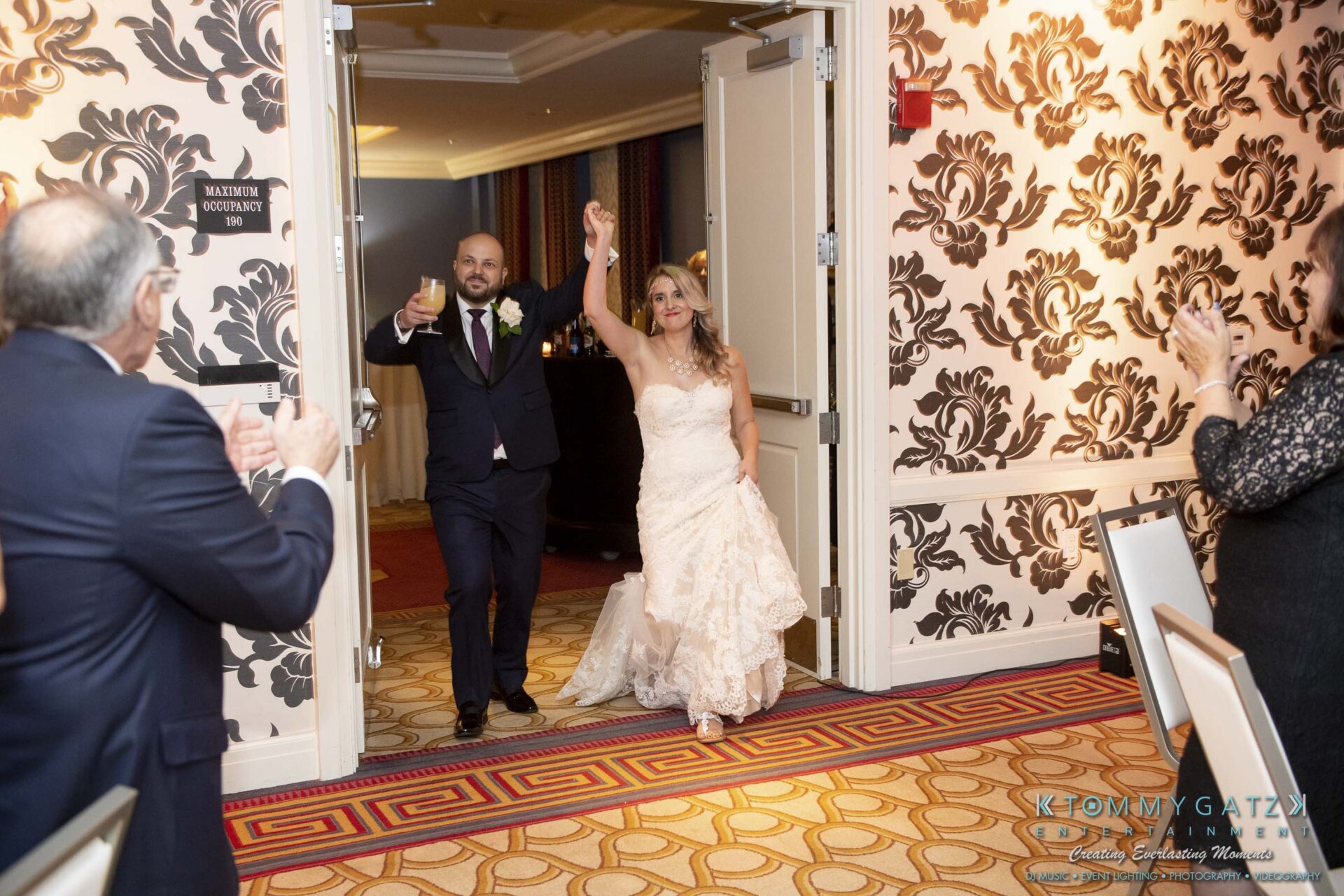 A bride and groom enter a ballroom holding hands, with guests applauding. The groom raises a glass, and the bride smiles. The room features patterned wallpaper and carpet.