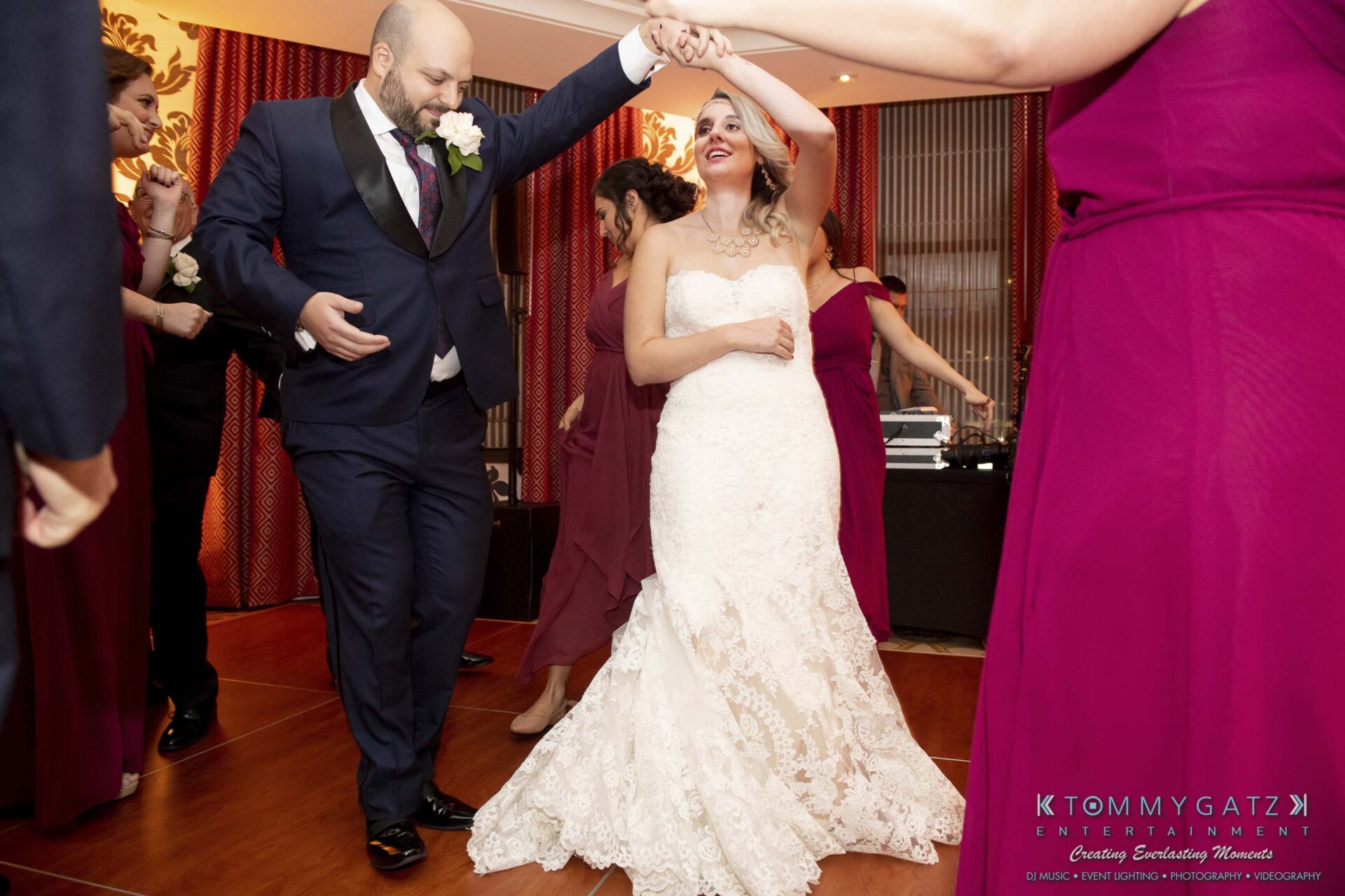A bride and groom dance joyfully on a wooden floor, surrounded by bridesmaids in burgundy dresses.