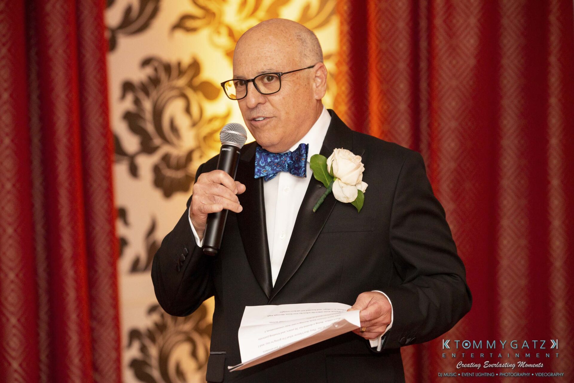 A man in a suit with a bow tie and white rose boutonniere speaks into a microphone while holding papers, standing against a decorative background.