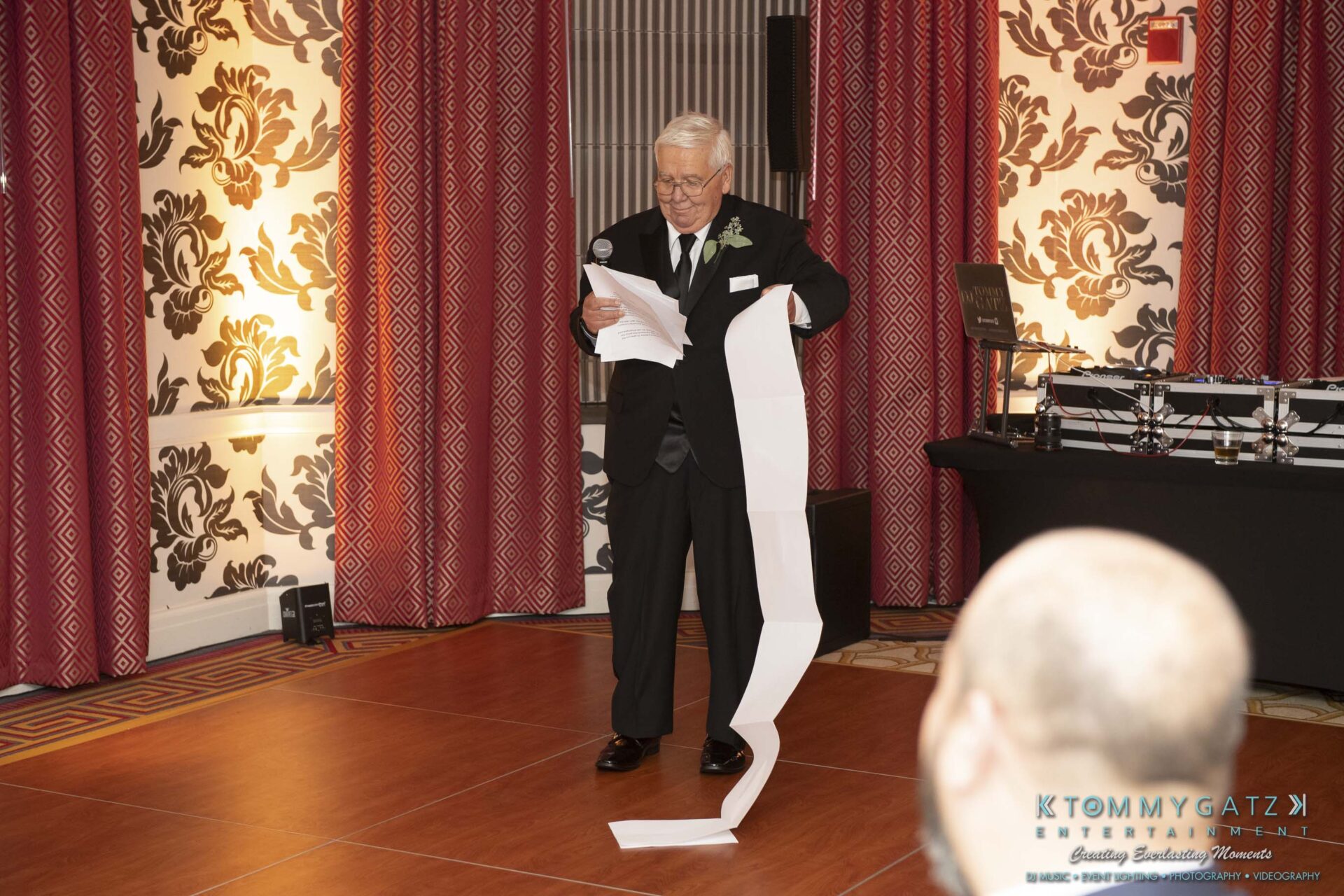 An elderly man in a suit reads from a long scroll of paper while holding a microphone, standing on a wooden floor with patterned curtains and a sound system in the background.