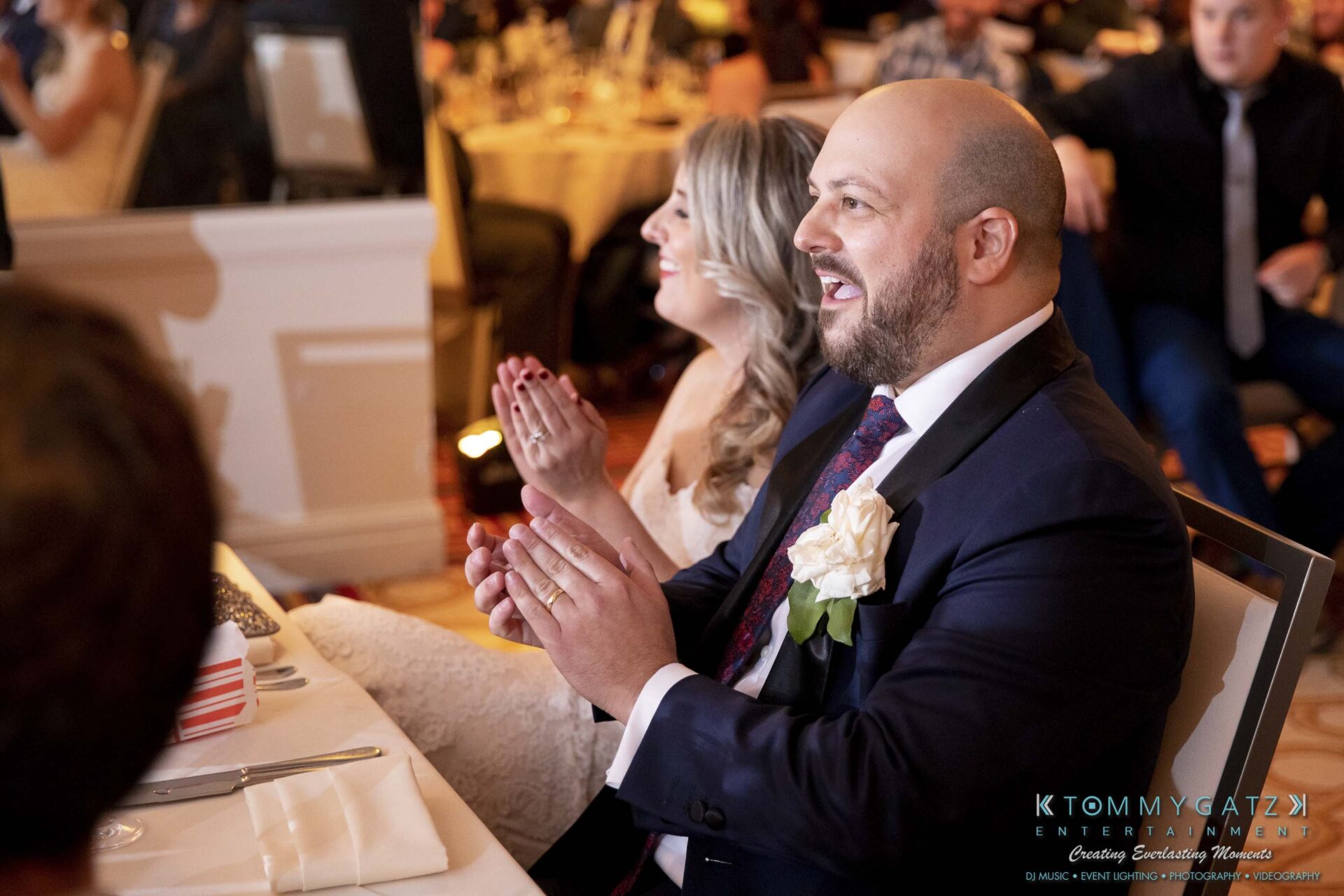 A man and woman are sitting at a table clapping, surrounded by other guests at an indoor event.