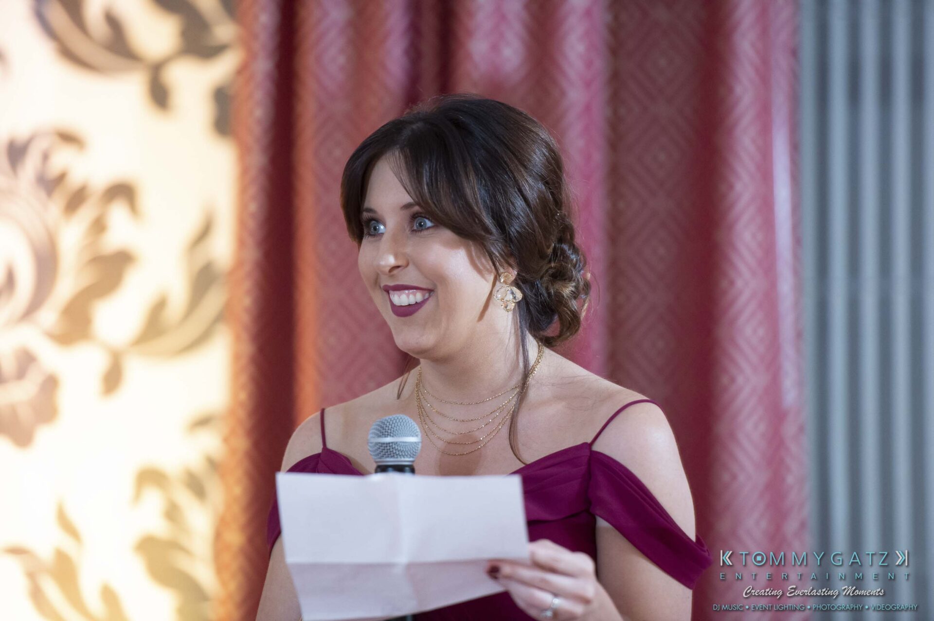 Woman in a purple dress holds a microphone and paper, smiling, in front of a patterned curtain.