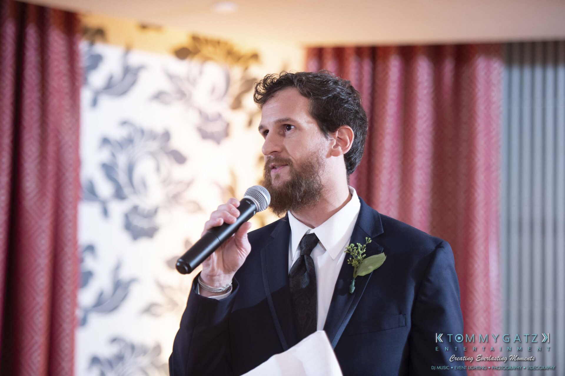 A man in a suit speaking into a microphone at an indoor event with floral wallpaper background.
