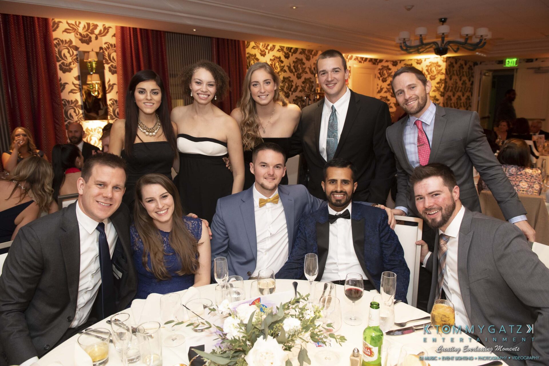 A group of people dressed formally, posing and smiling around a dinner table with plates, drinks, and a centerpiece in a warmly decorated room.