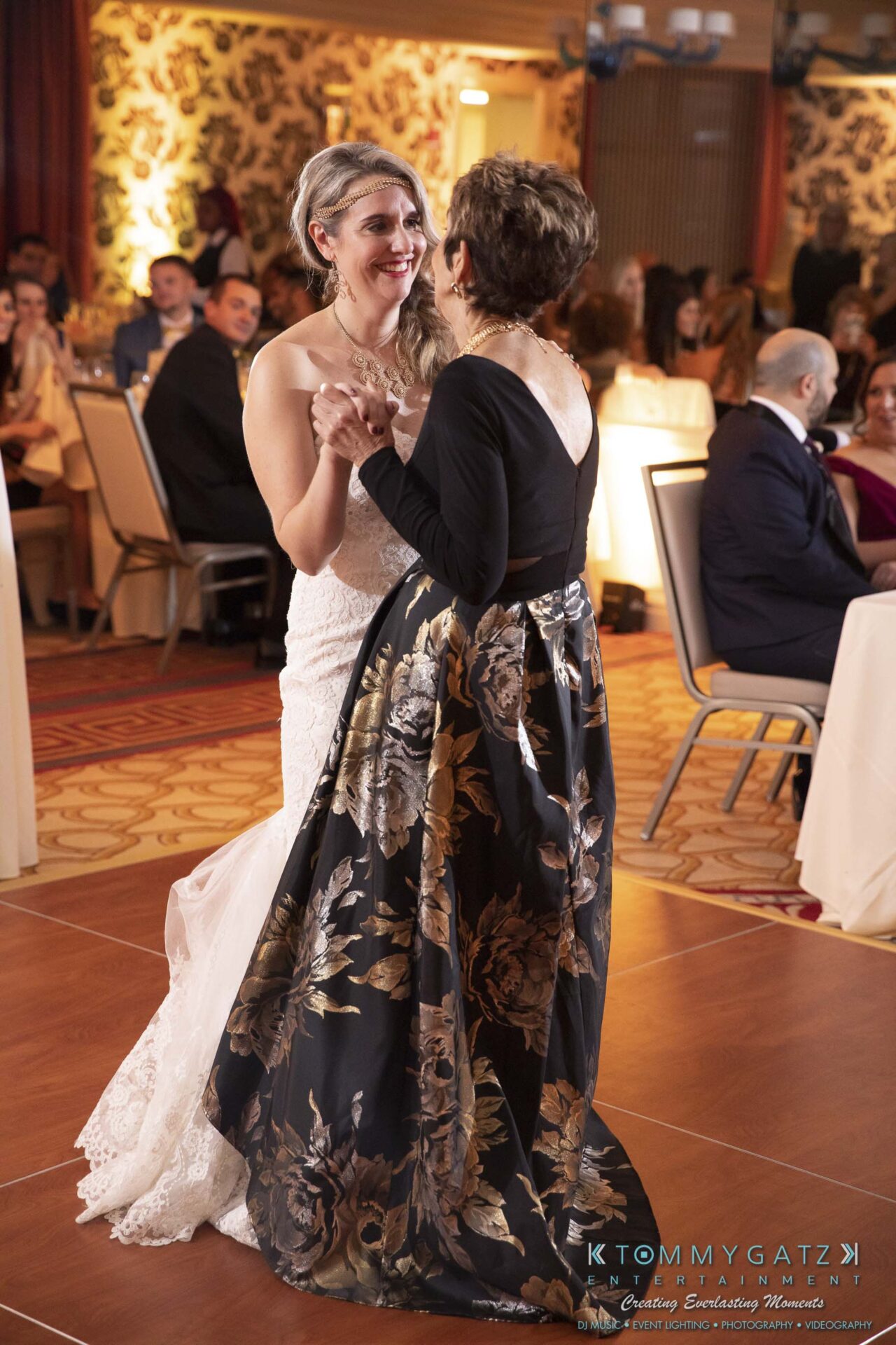 Two women dancing together at a formal event. One is in a wedding dress and the other in a floral gown. Guests are seated in the background.